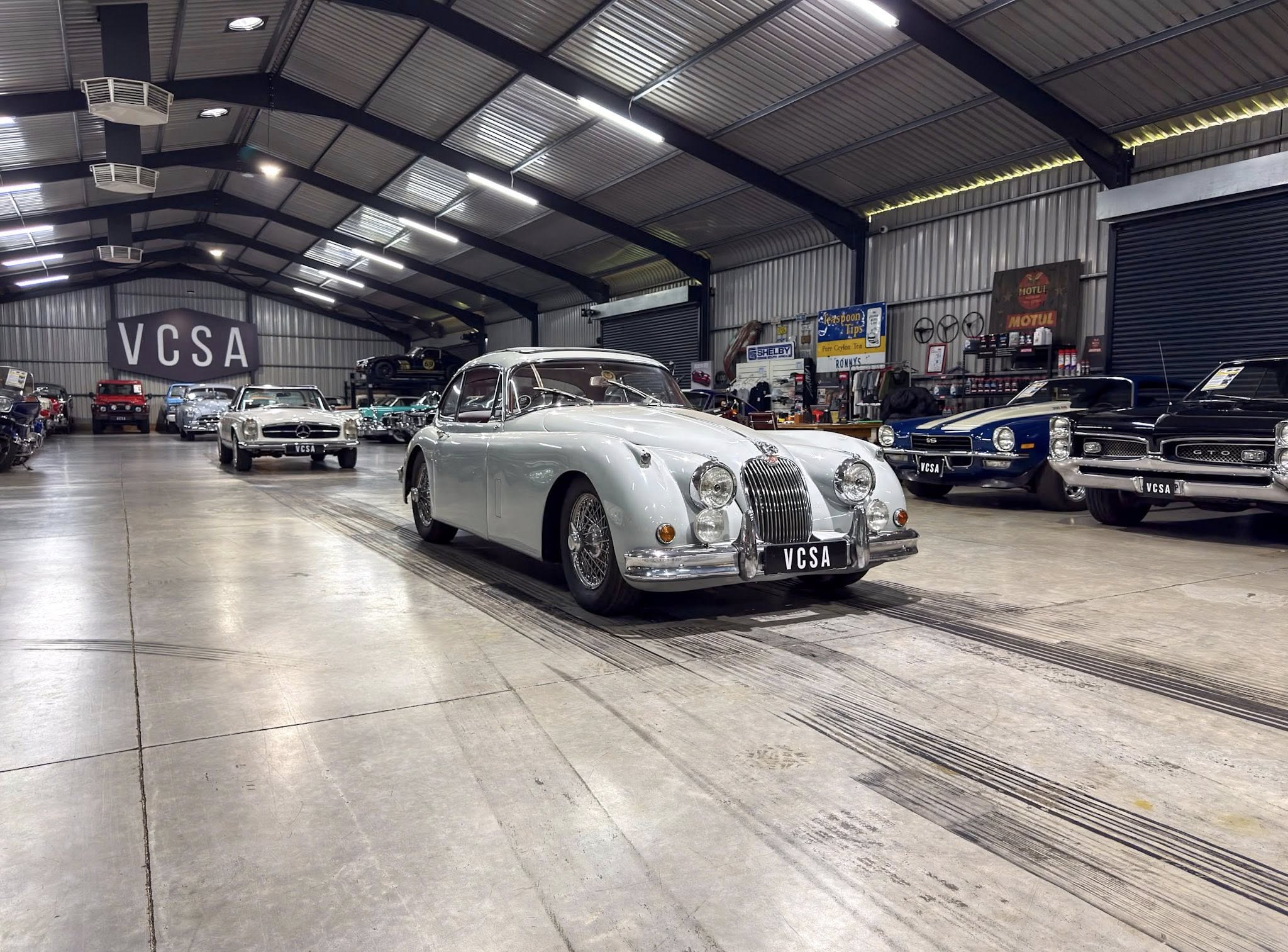 Indoor showroom with vintage cars lined up inside a large industrial warehouse; prominent 'VCSA' sign on wall.