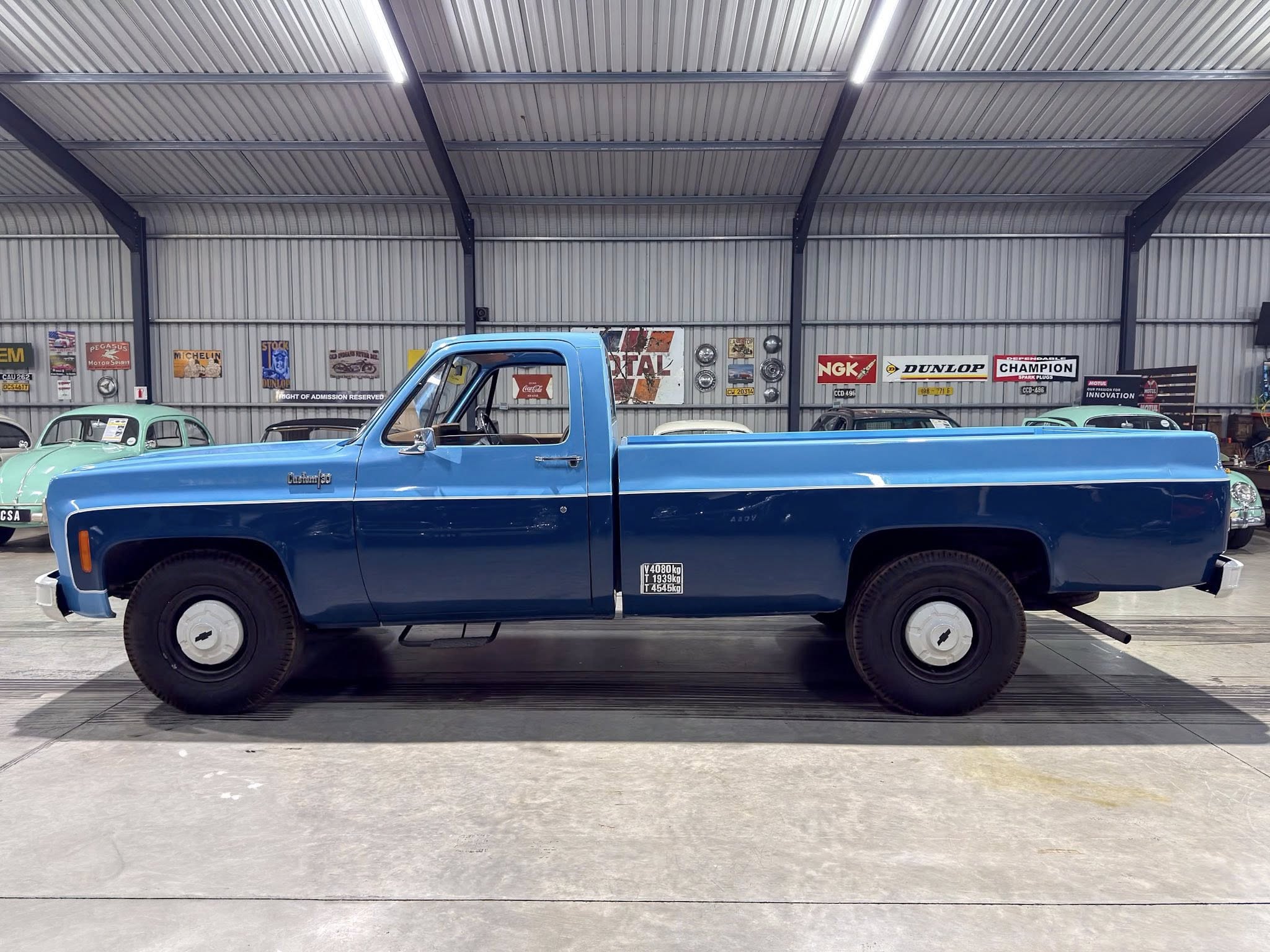 Blue vintage pickup truck parked inside a metal-walled showroom with retro signs on the wall behind it.