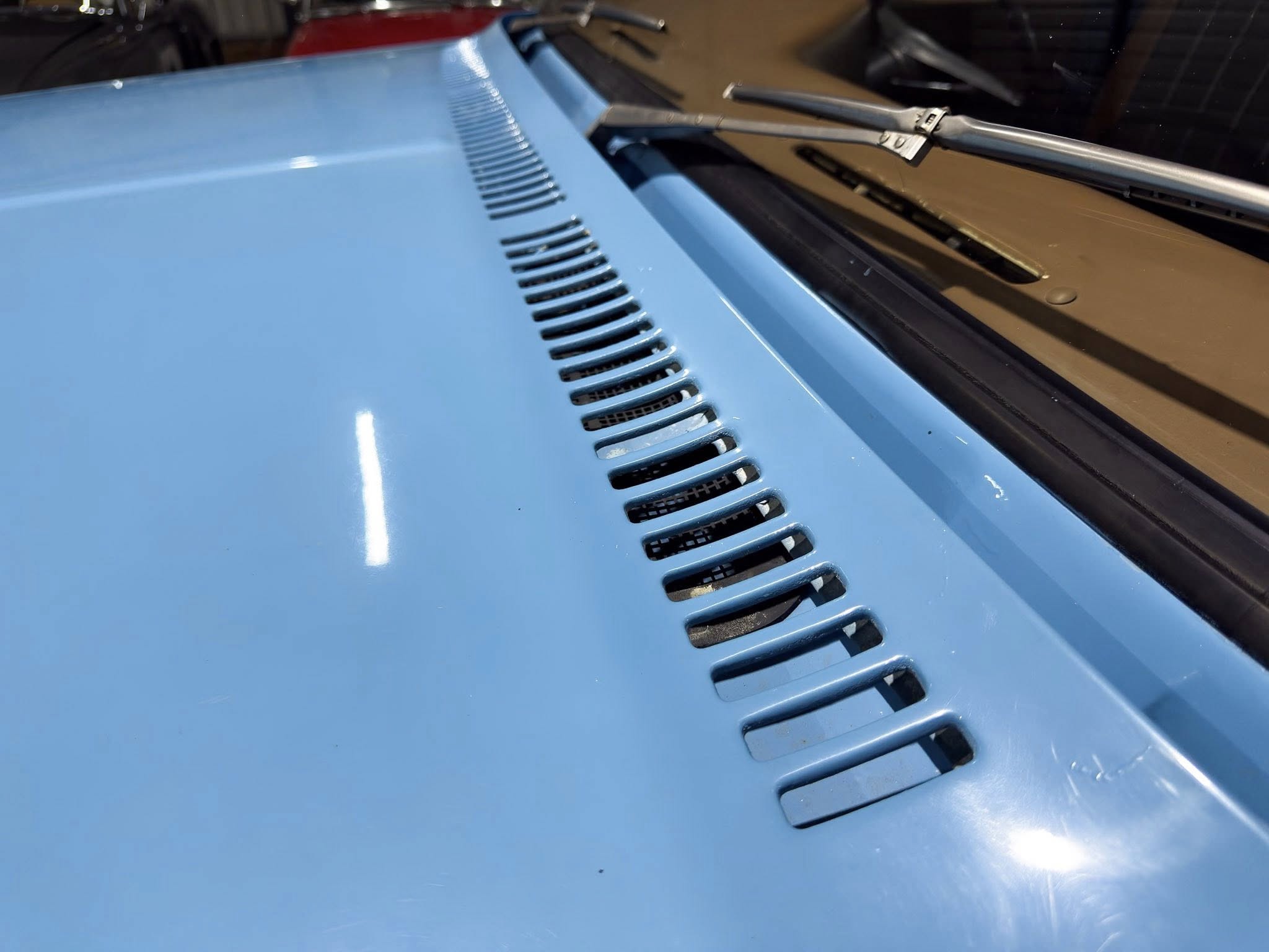 Close-up of a blue car hood with vent slits and a pair of windshield wipers resting on the windshield edge, taken at an angle.