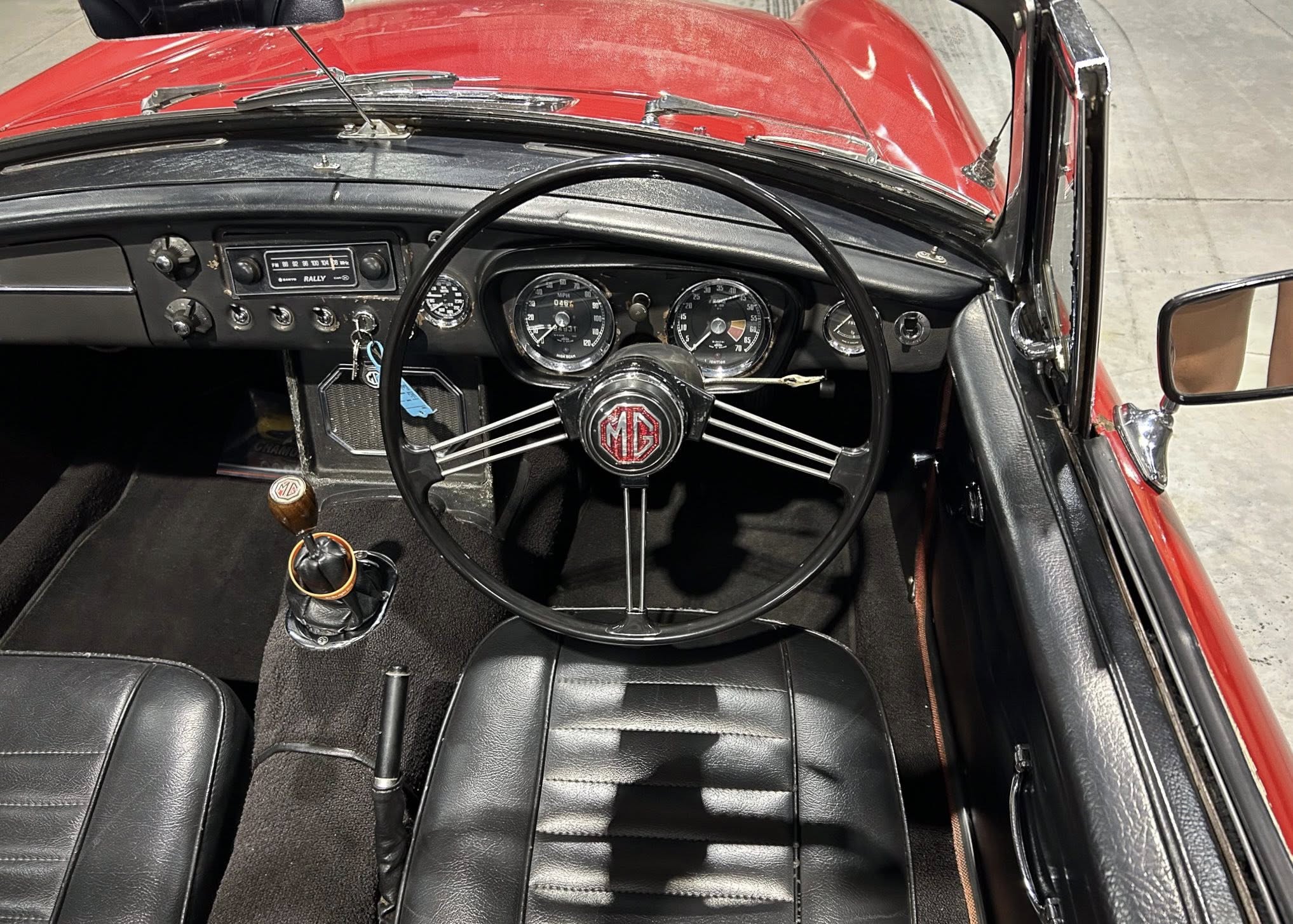 View from the driver’s seat of a red vintage MG sports car: black dashboard, large round gauges, and a classic three‑spoke steering wheel with MG emblem, with red exterior visible through the windshield.