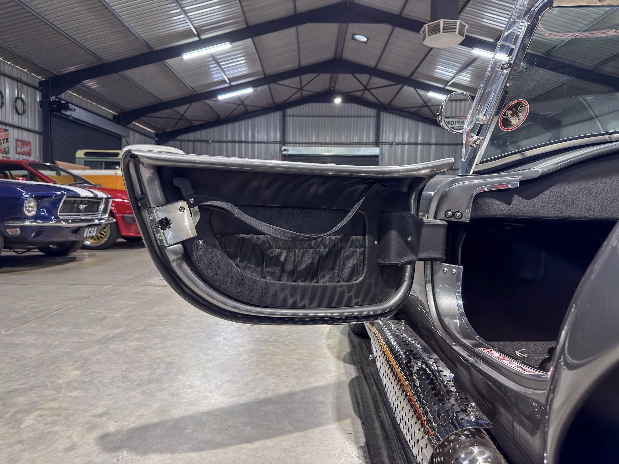 Open car door of a vintage black convertible inside a spacious metal workshop with other classic cars in the background.