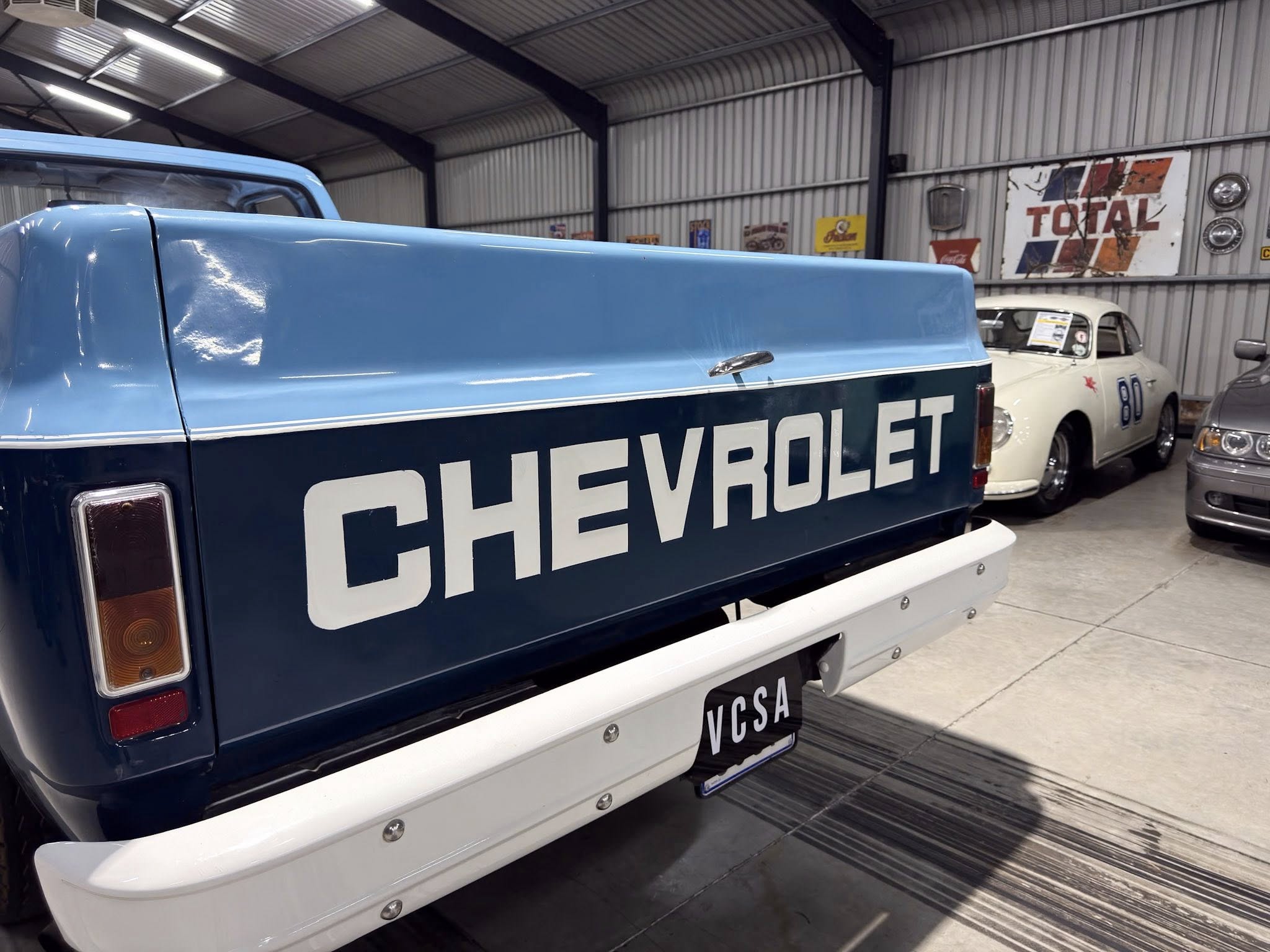Blue Chevrolet pickup tailgate in a showroom with vintage cars and a Total sign on the wall in the background.