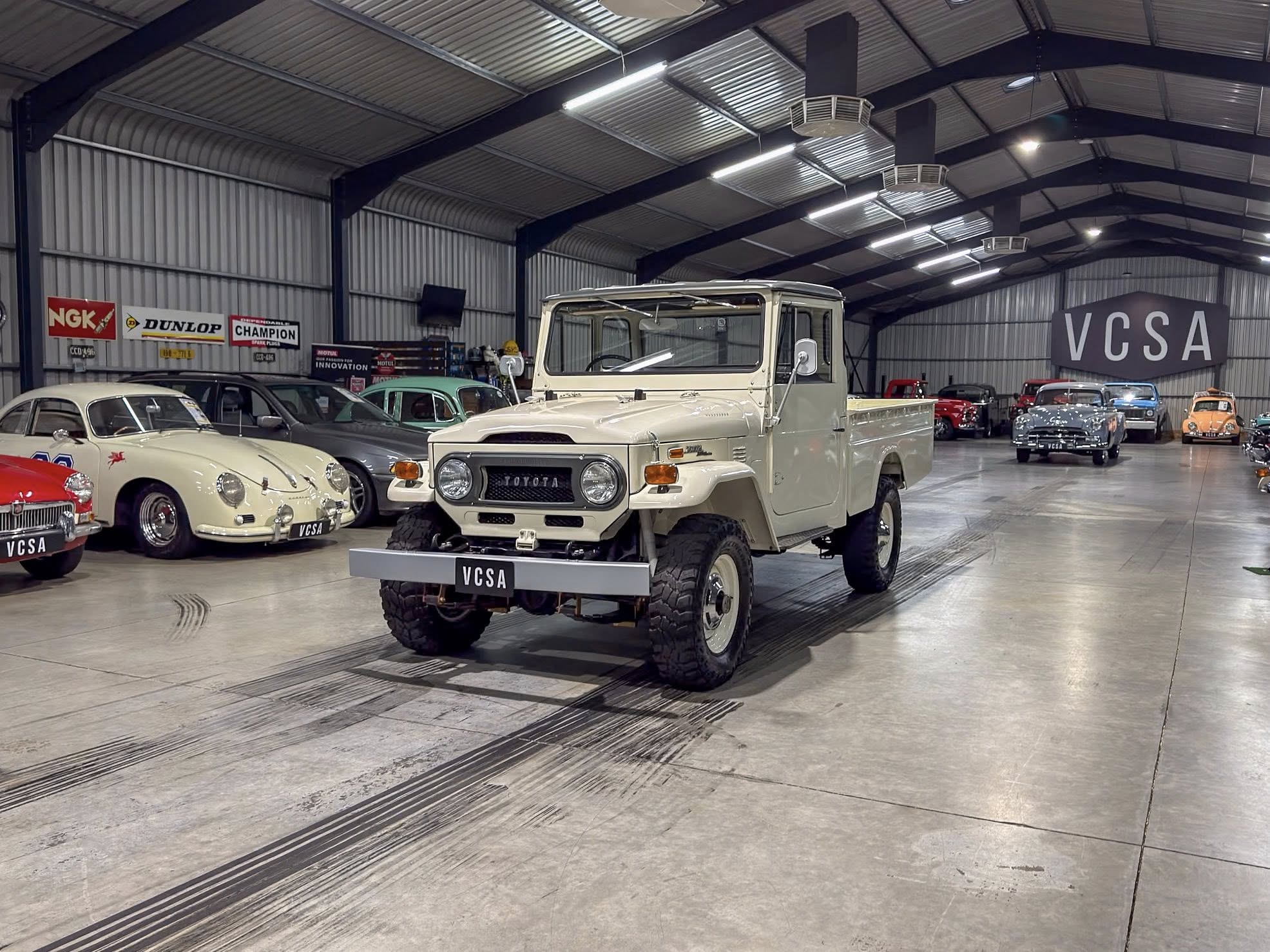 Indoor showroom of vintage cars, foreground features a beige Toyota pickup with other classic cars nearby in a large, well-lit space: VCSA signage visible in back.