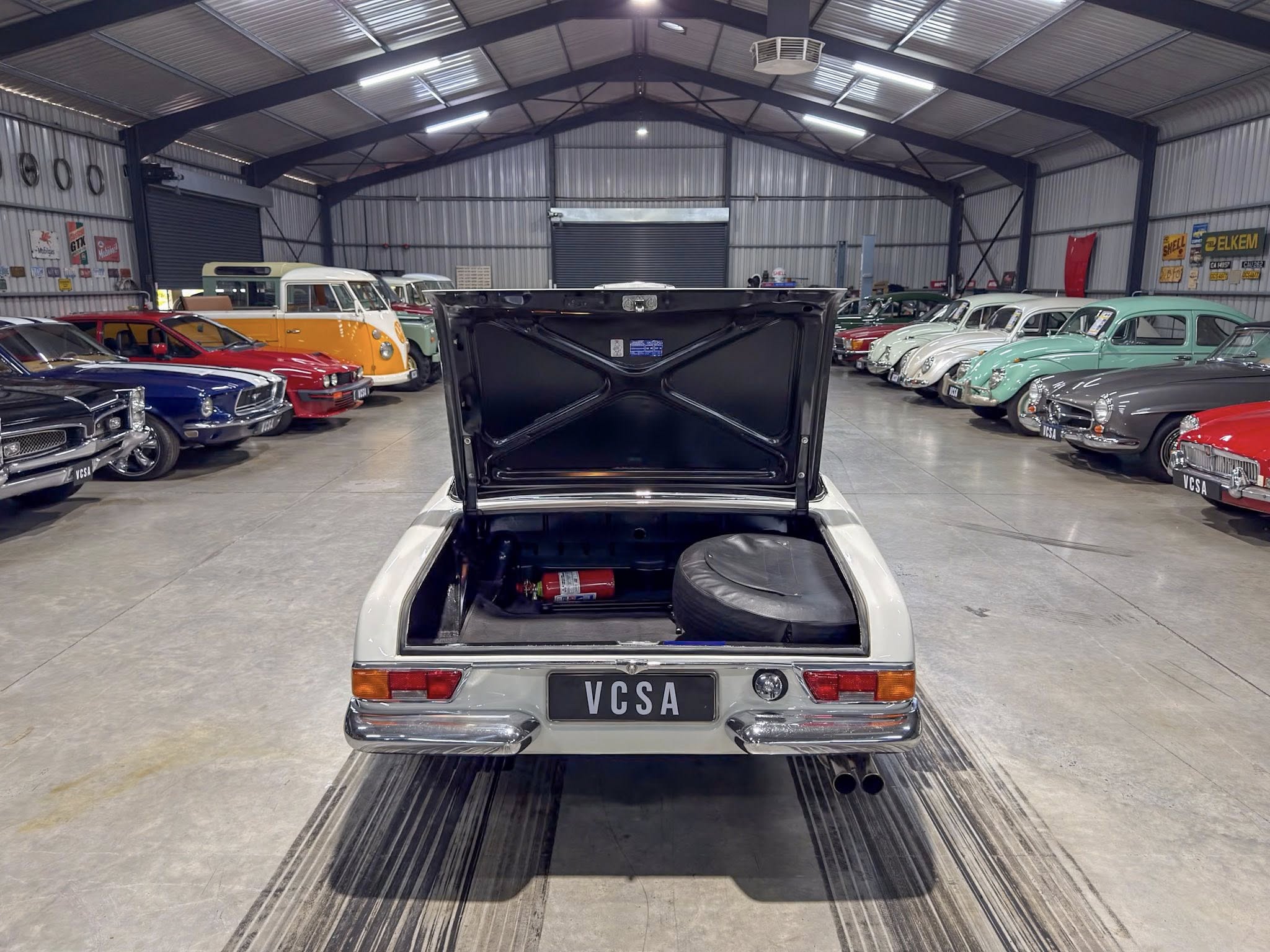 White vintage car with its trunk/hood open seen from behind, inside a large showroom filled with colorful classic cars lined up on both sides.