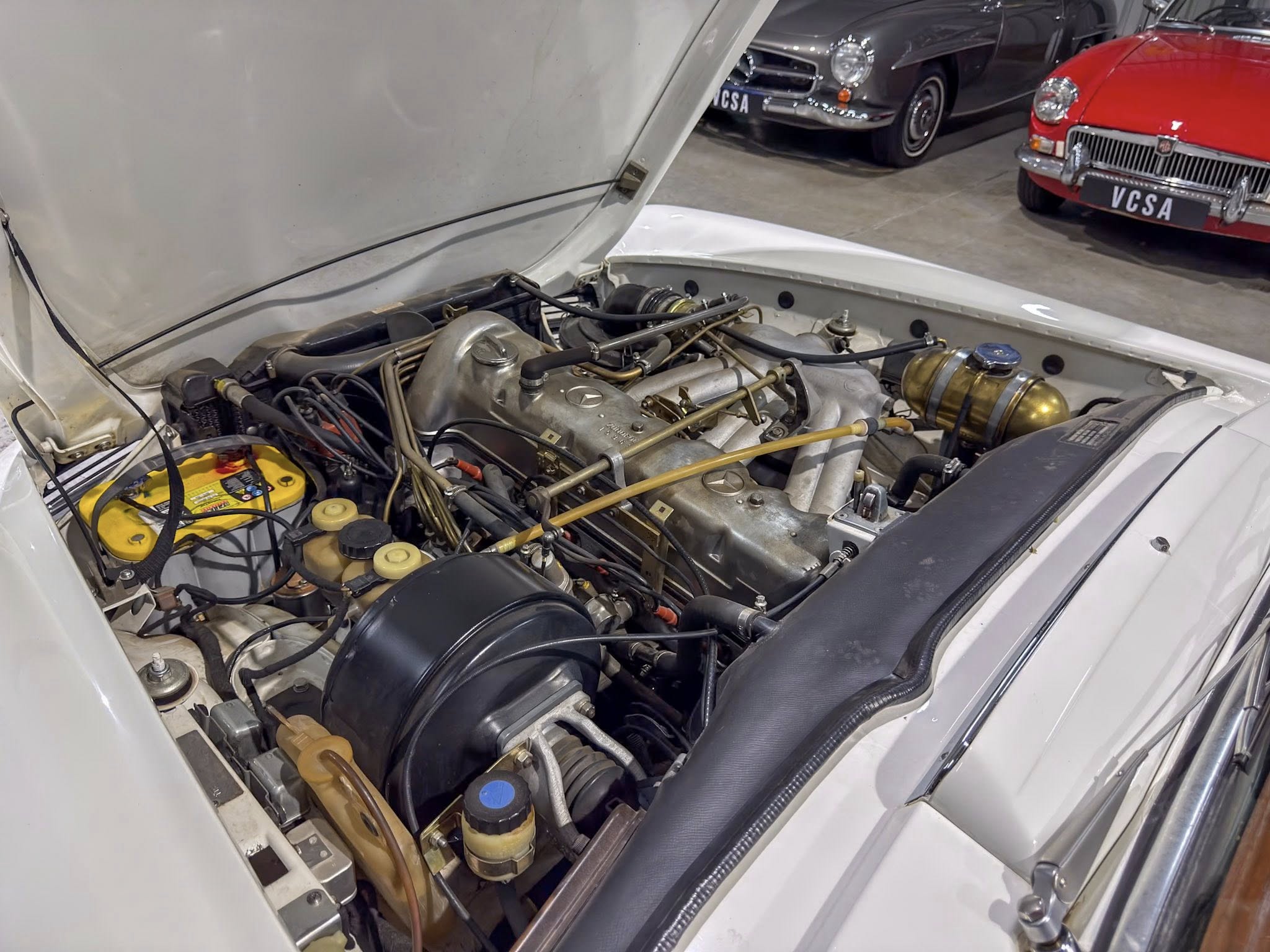 Open hood of a classic car showing the engine bay with hoses and metal components, nearby vintage cars on display in a showroom.