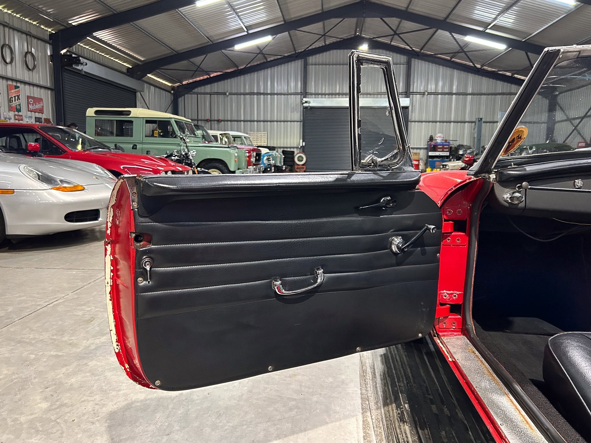 Inside a classic car workshop with a red interior door open; green vintage van and other old cars in the background.