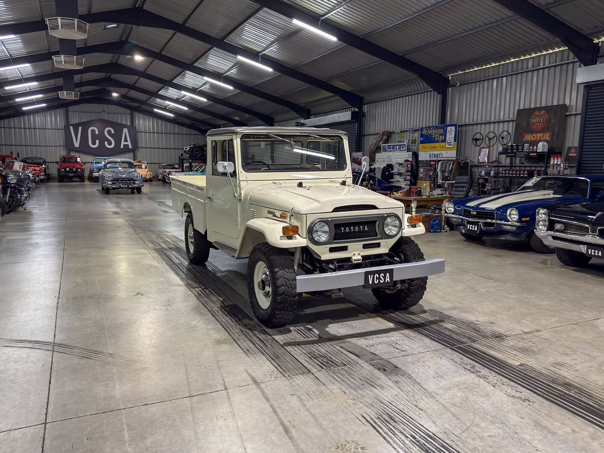 Indoor showroom filled with classic cars, a white Toyota pickup in the foreground and a large VCSA sign in the background.
