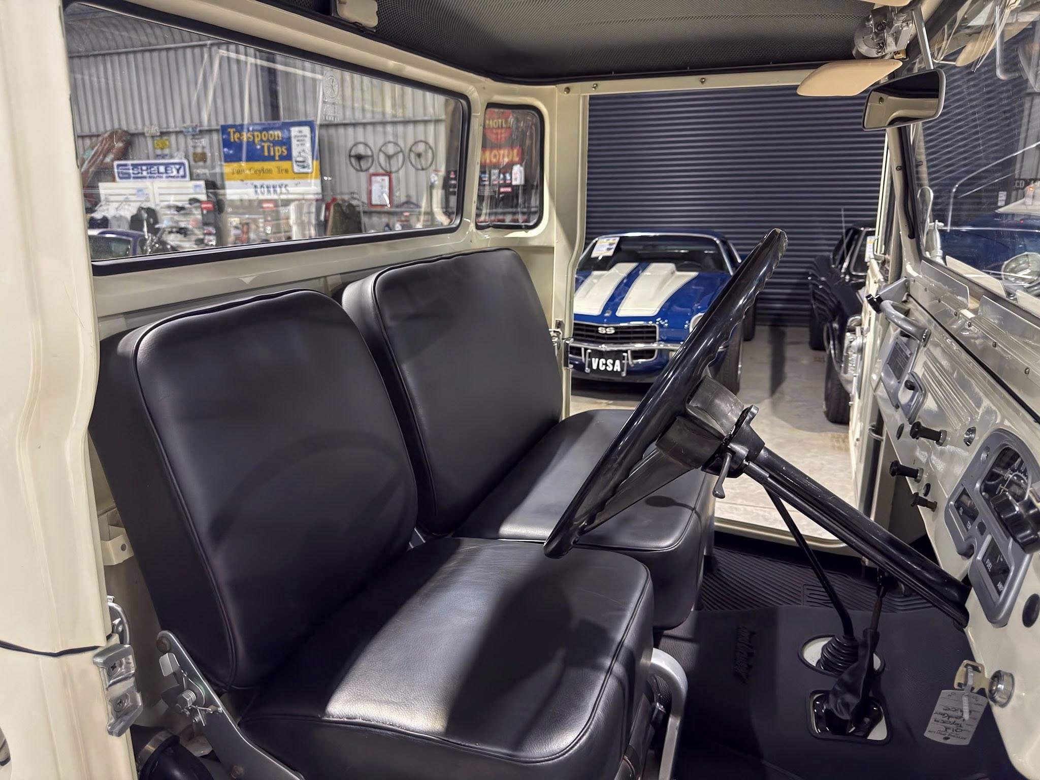 Interior of a vintage utility vehicle showing black vinyl seats, steering column, and metal dashboard, with a blue car visible through the open window in a showroom.