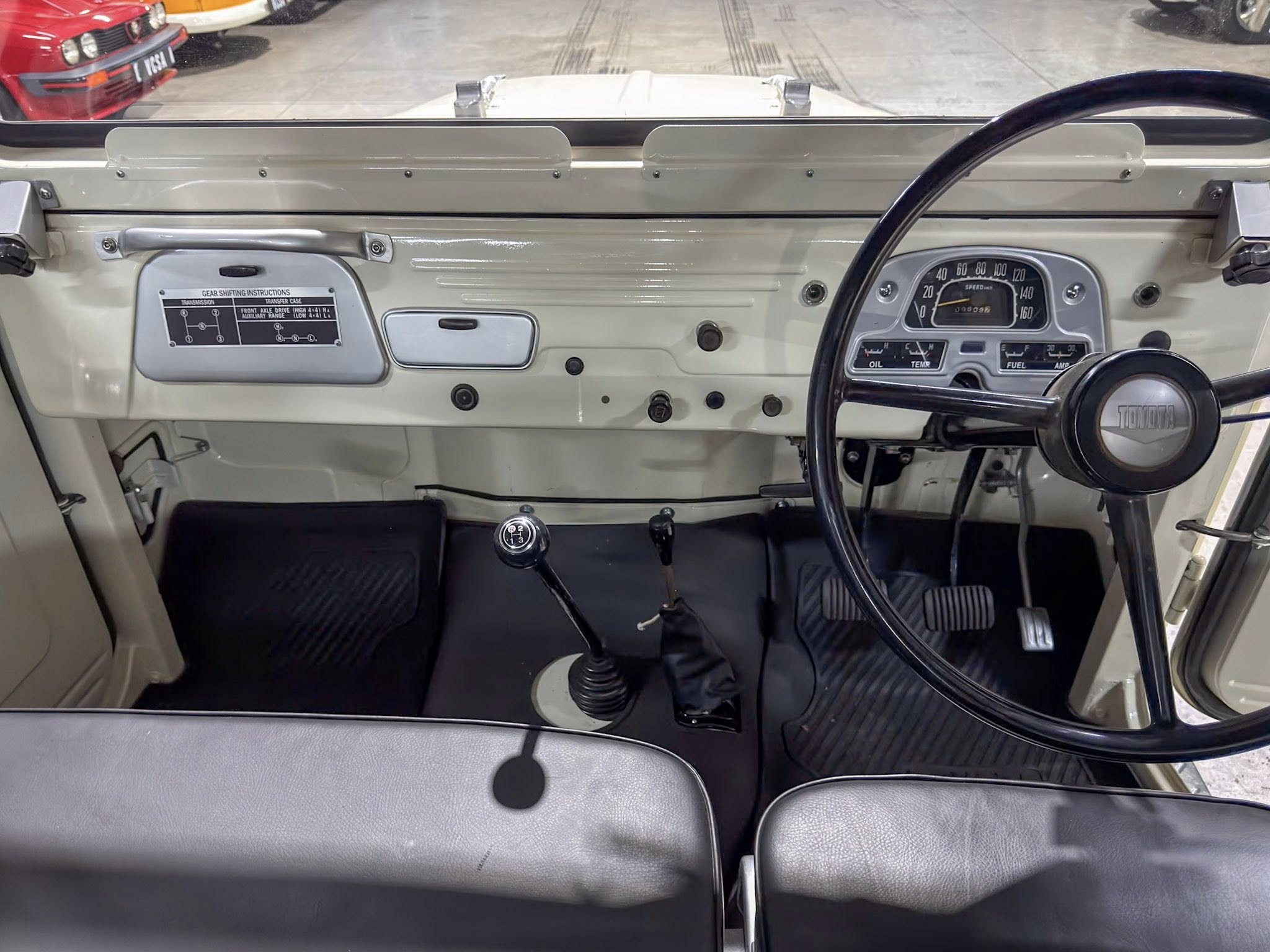 Vintage car dashboard interior with a right-hand steering wheel, classic gauges, and a floor-mounted gear lever seen from the passenger seat area.