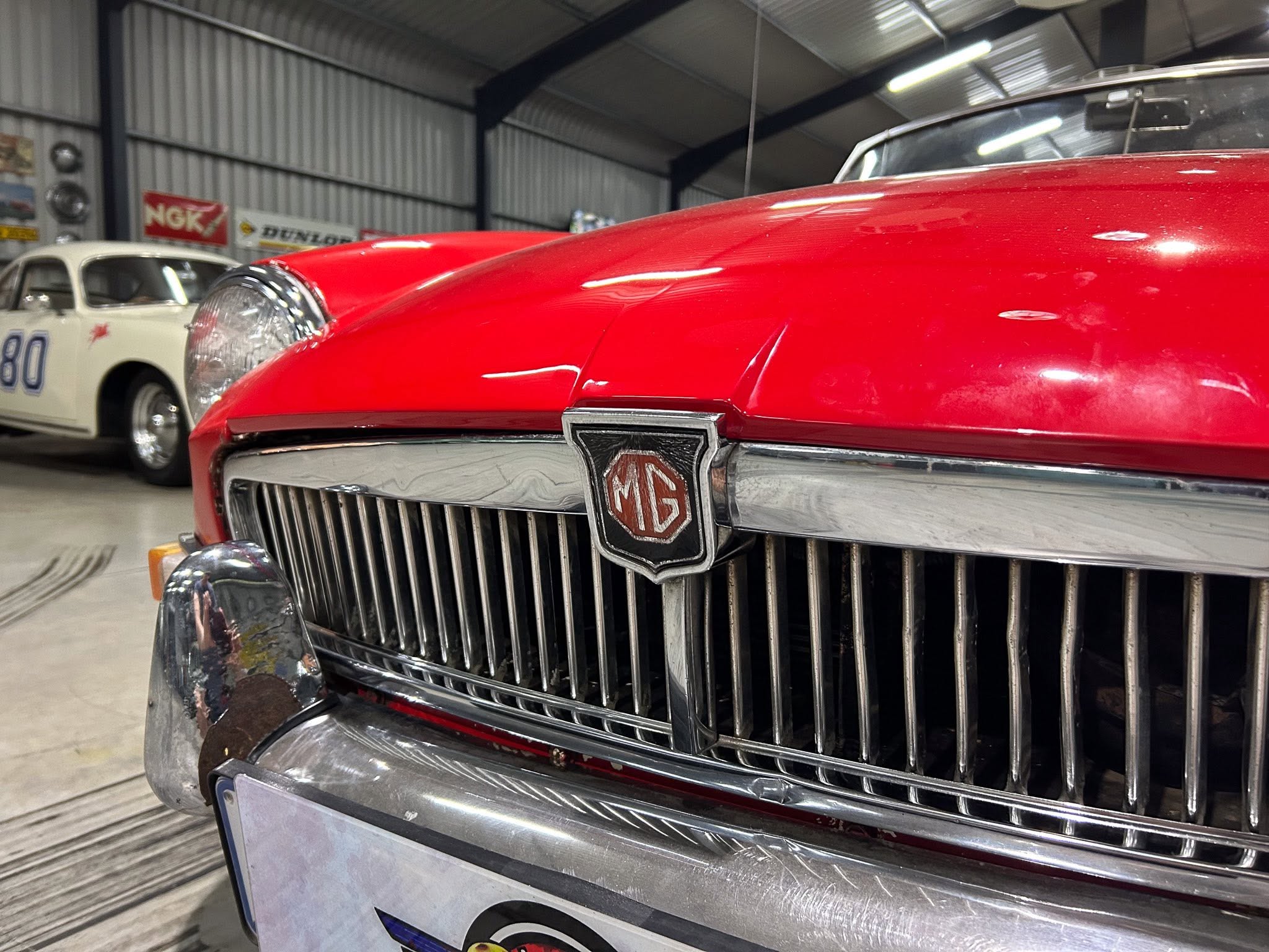 Close-up of a red MG car grille with chrome bumper, displayed in a vintage showroom with other classics in the background.