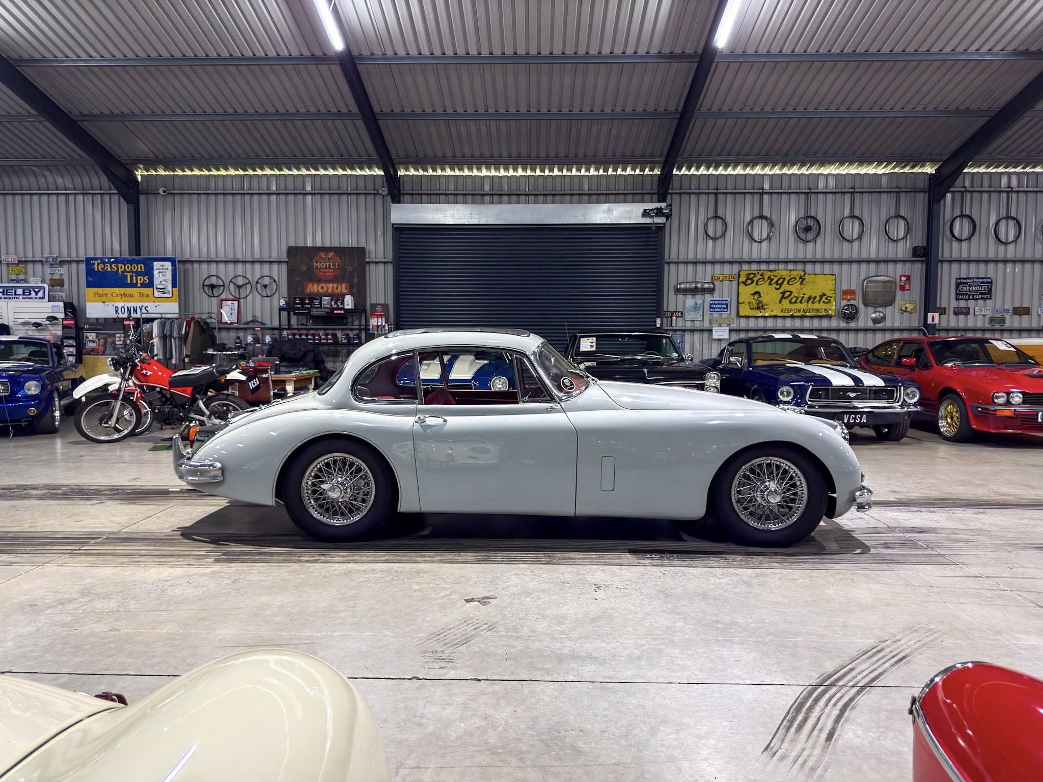 Powder-blue vintage car in the foreground inside a metal-walled showroom filled with classic cars and retro signage along the walls.
