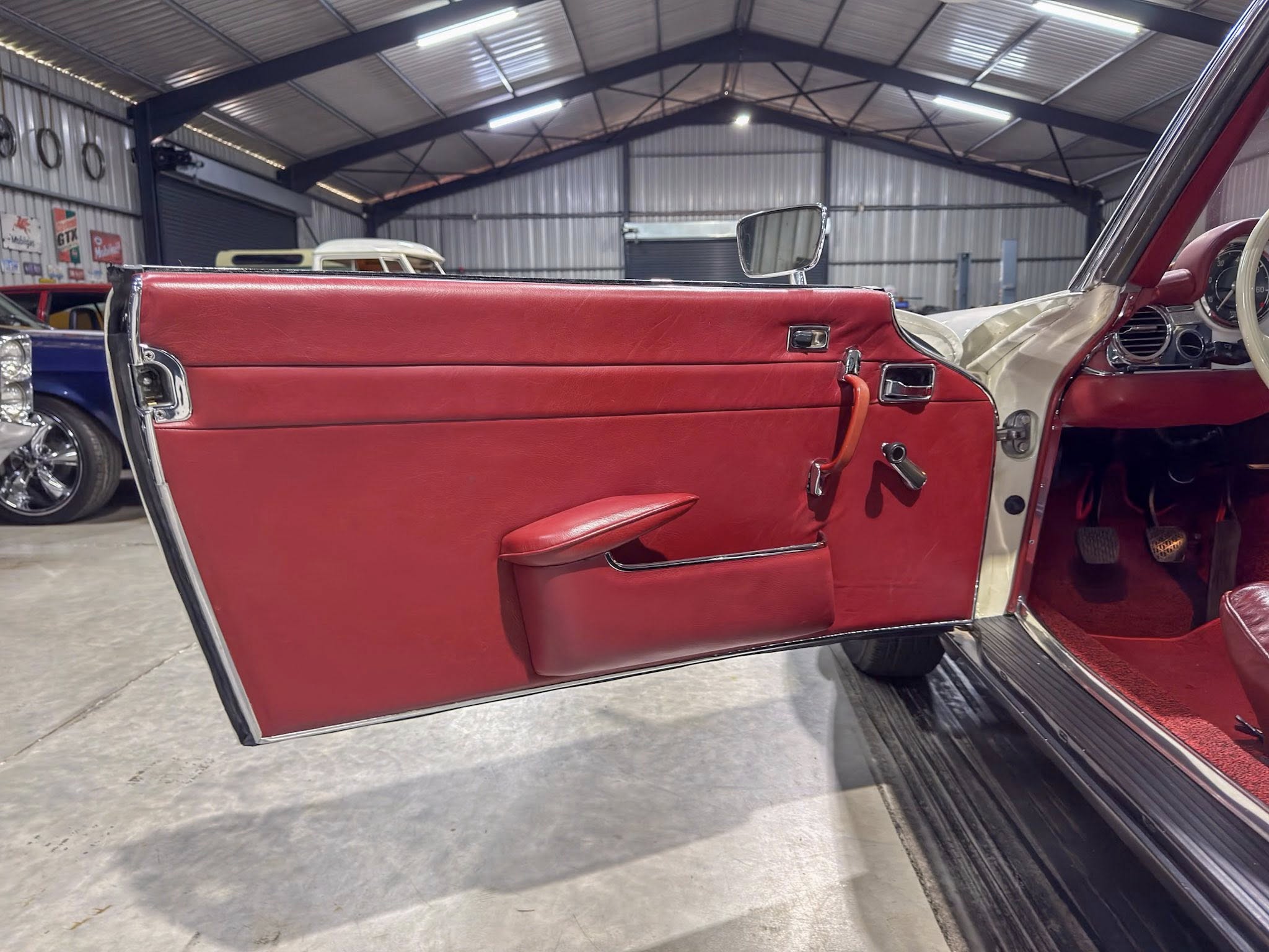 Open red car door inside a classic car showroom with chrome trim and red leather interior; other cars visible in the background, metal warehouse walls behind.