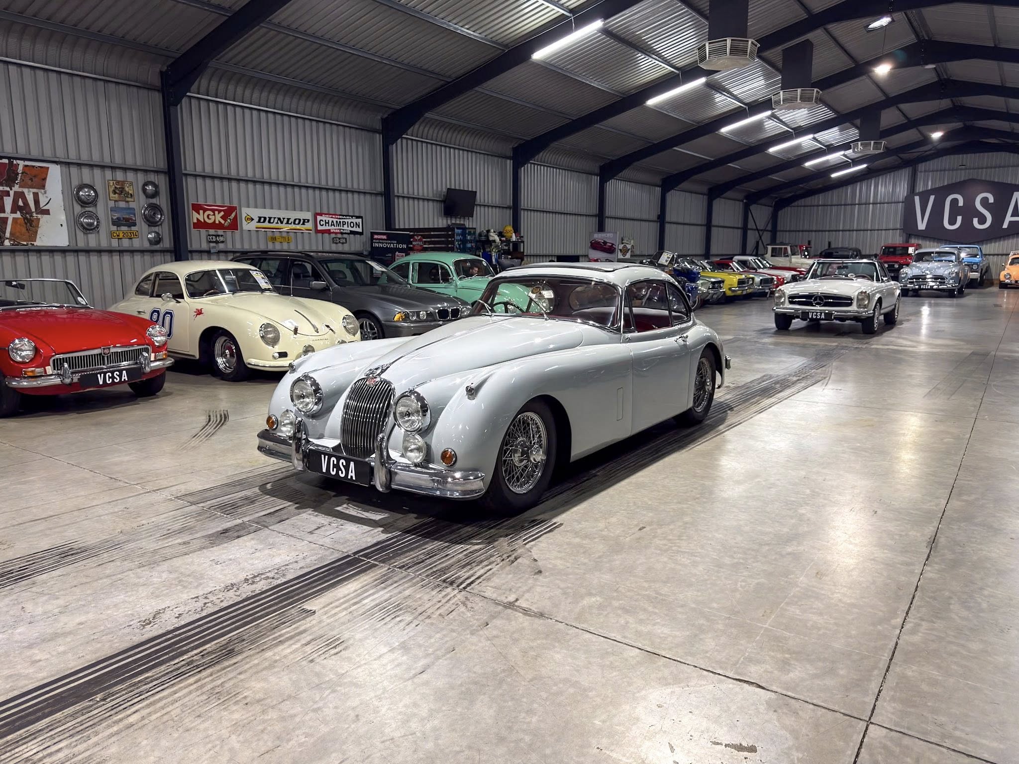 Interior of a large showroom with a line of classic cars, including a silver vintage convertible in the foreground and a red car at left (VCSA license plates).