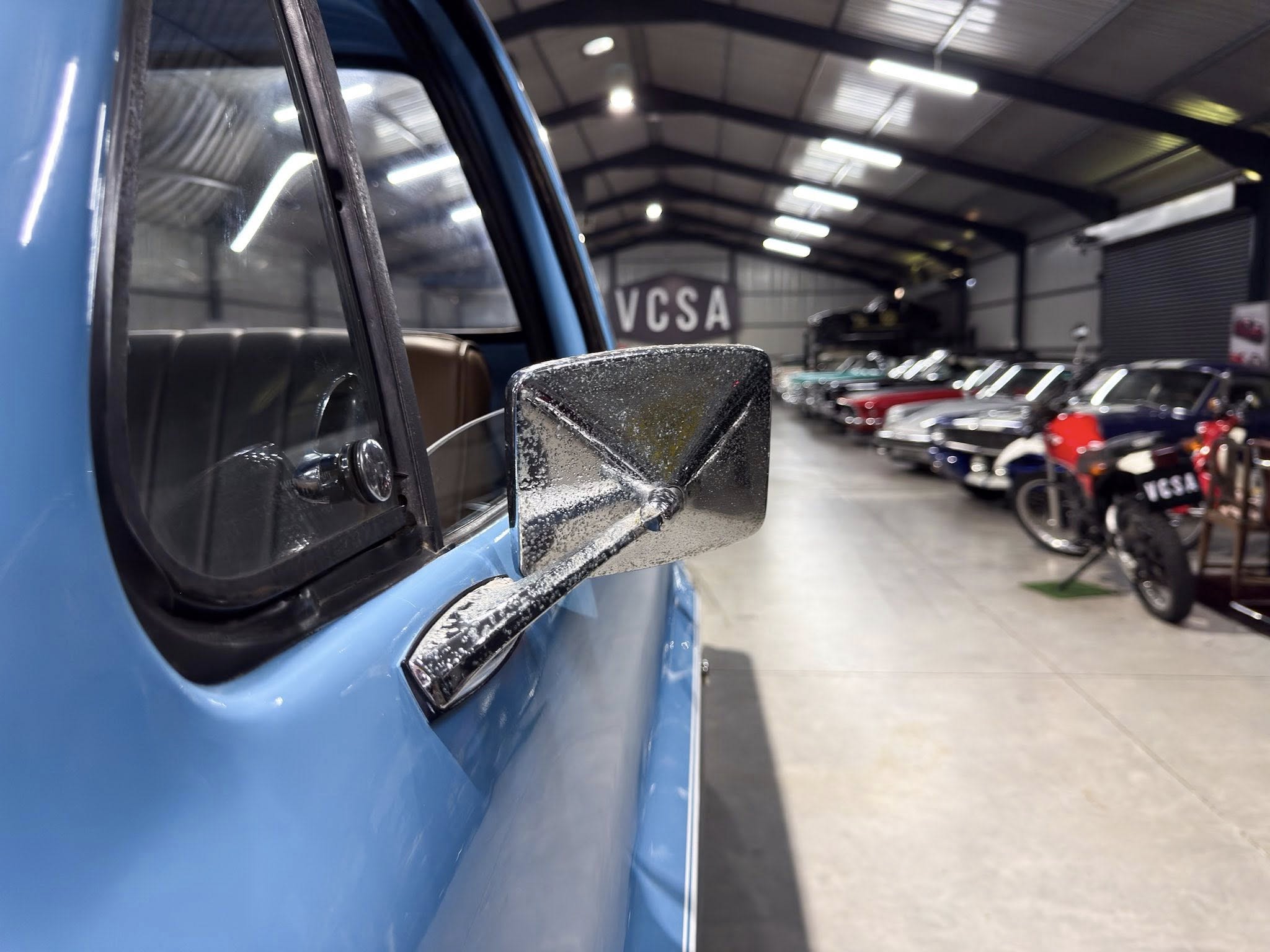 Close-up of a blue car's side mirror and window in a showroom or garage. Nearby cars line the background.
