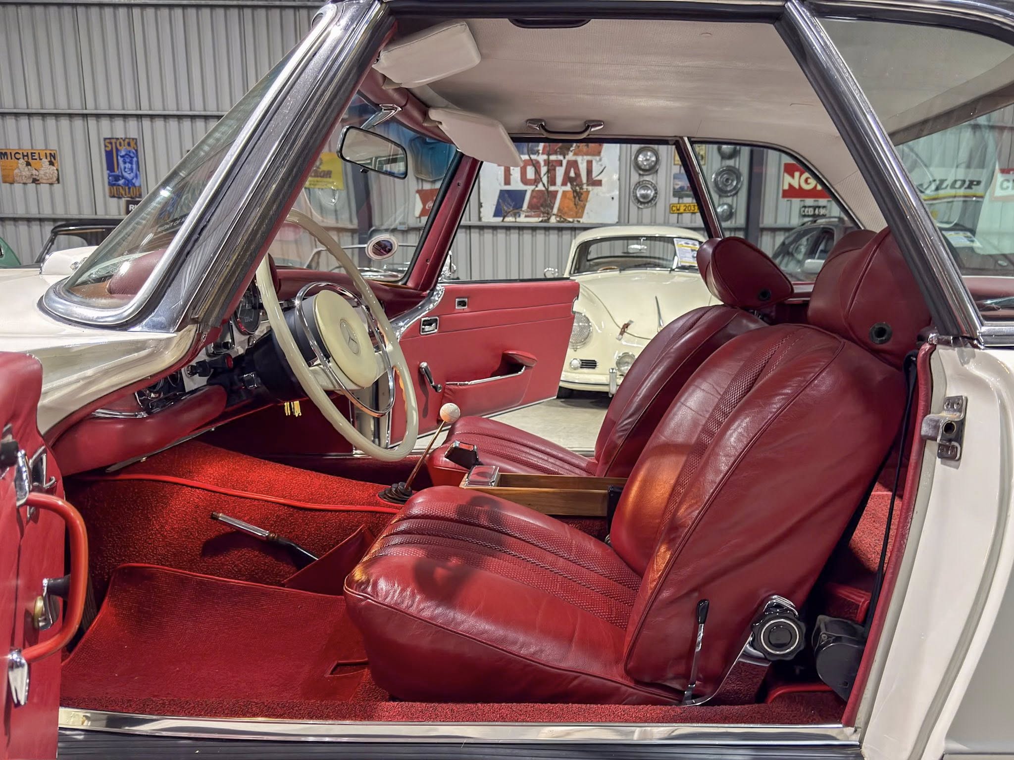 Vintage car interior with red leather seats, cream steering wheel, and chrome detailing seen through an open door in a showroom.