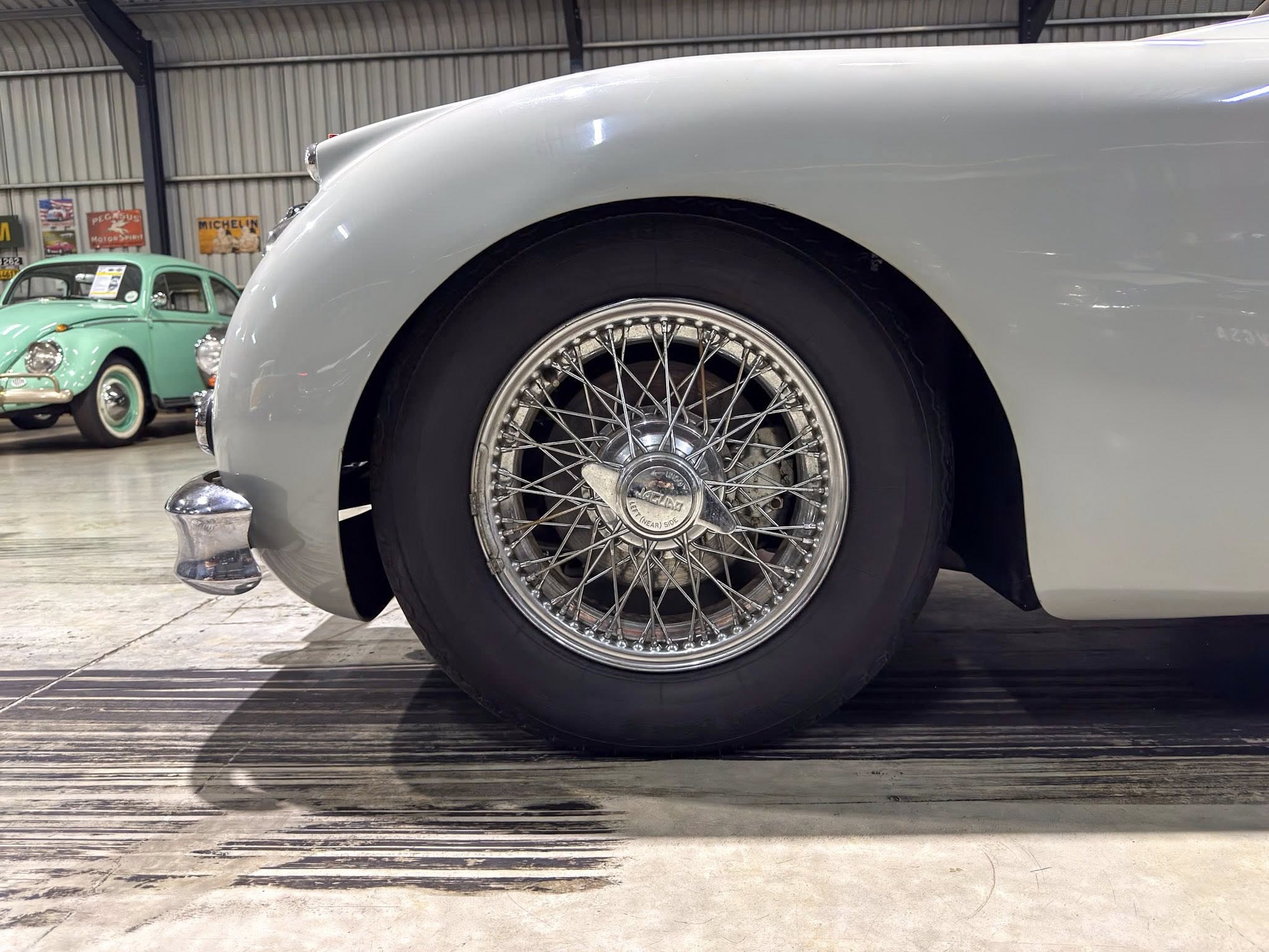 Close-up of a vintage car wheel with wire spokes on a light gray car in a showroom, background shows a mint green classic car.