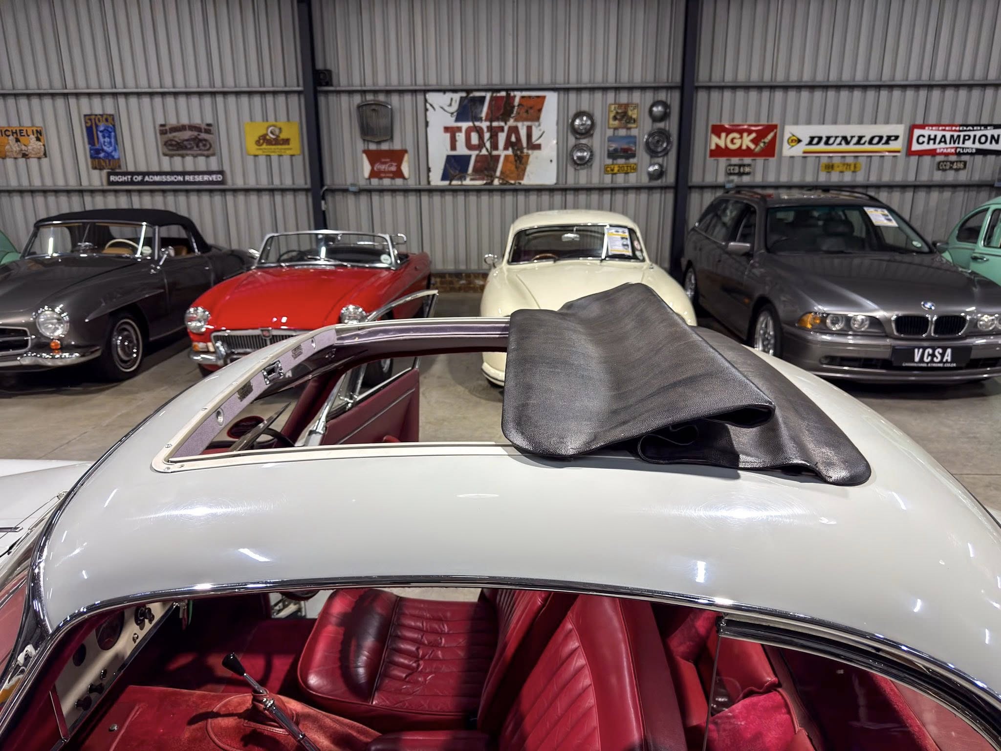 Open-top white vintage car in foreground inside a showroom, red leather seats visible, with classic cars and retro signs along corrugated metal wall behind it.