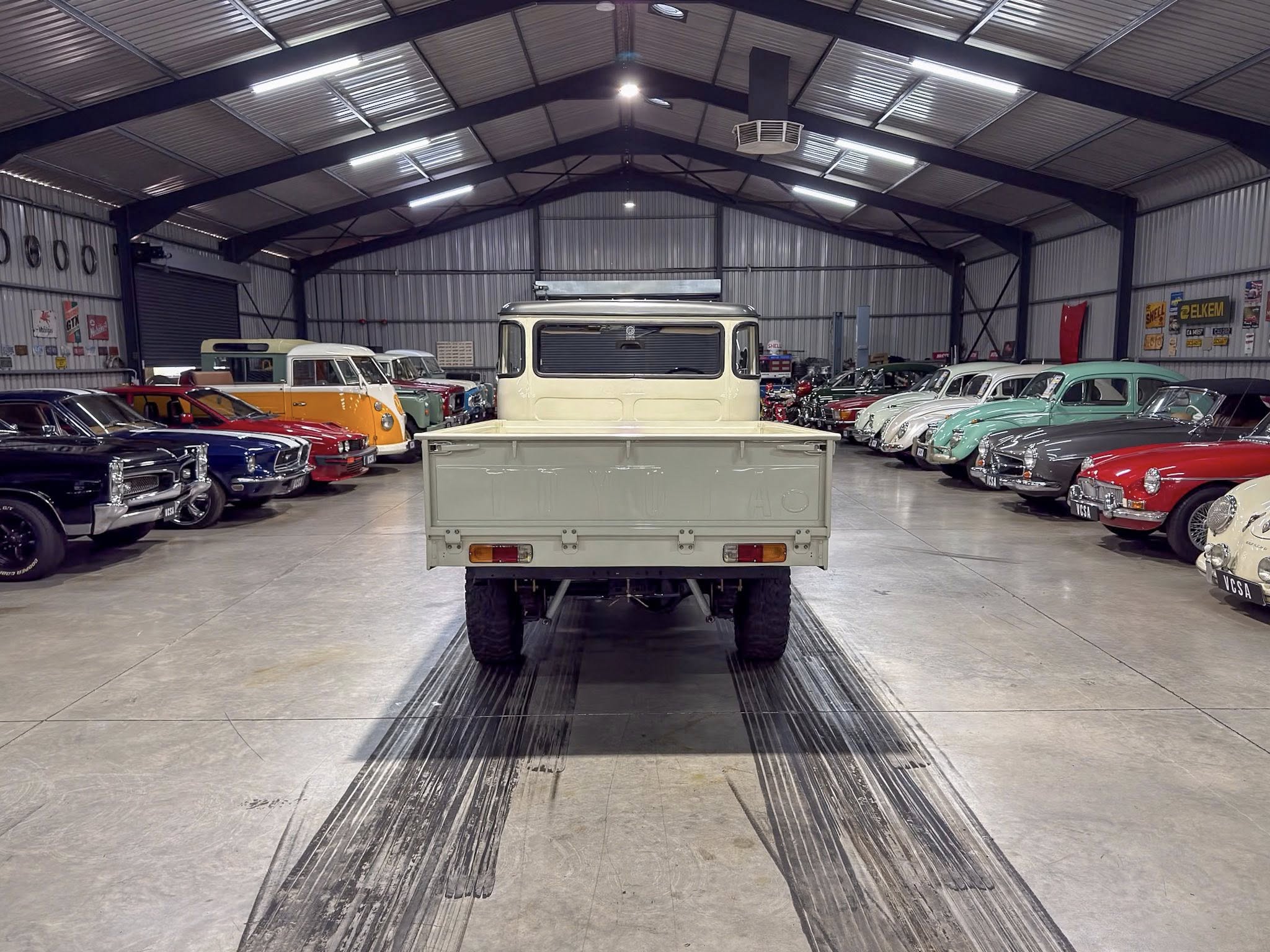 Interior of a large, metal-walled warehouse filled with classic cars, beige pickup centered in the foreground.