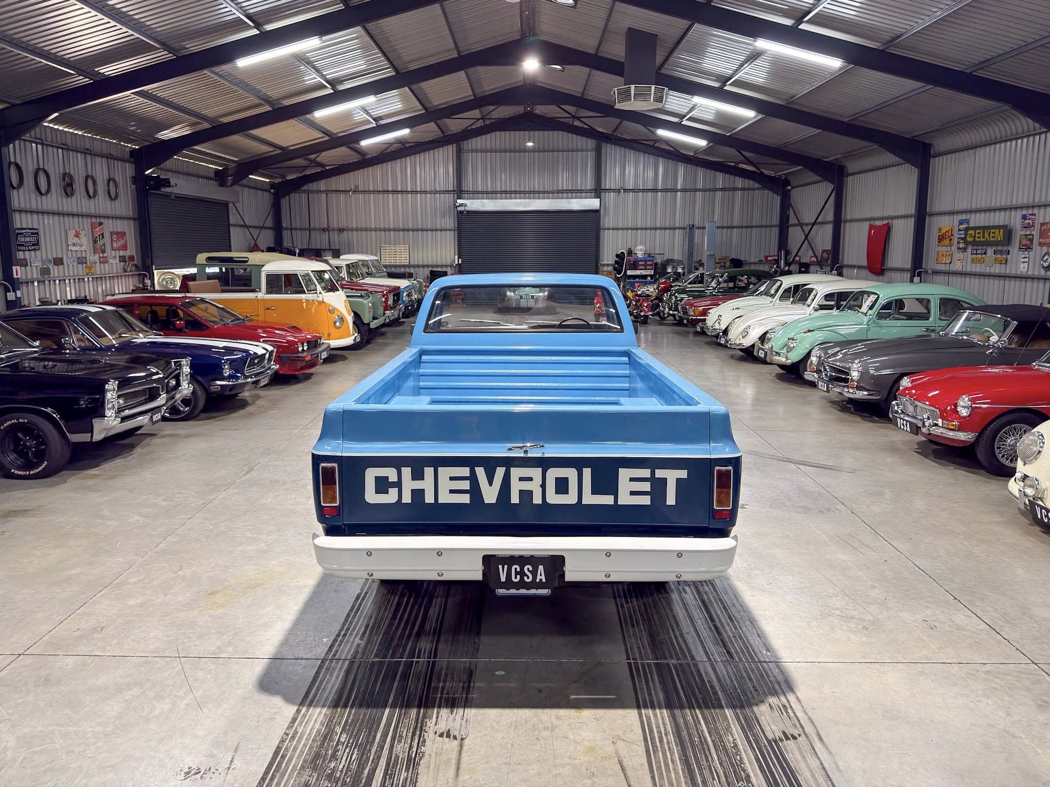 Rear view of a light blue Chevrolet pickup in a vintage car showroom with rows of classic cars along both sides and a Chevrolet sign on the tailgate