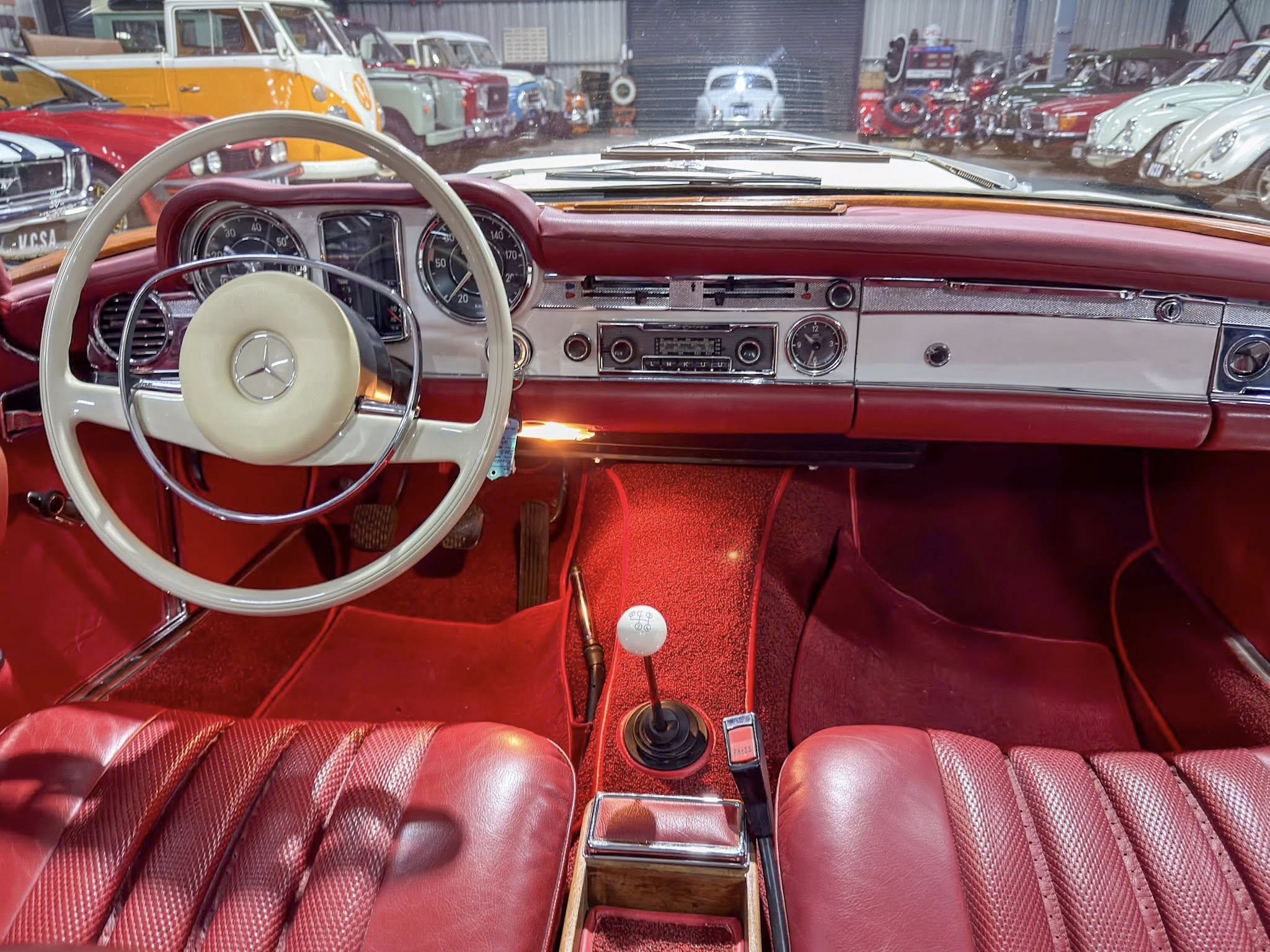 Red vintage car interior with cream steering wheel, chrome dashboard and classic gauges; other classic cars visible in a showroom outside the windshield.
