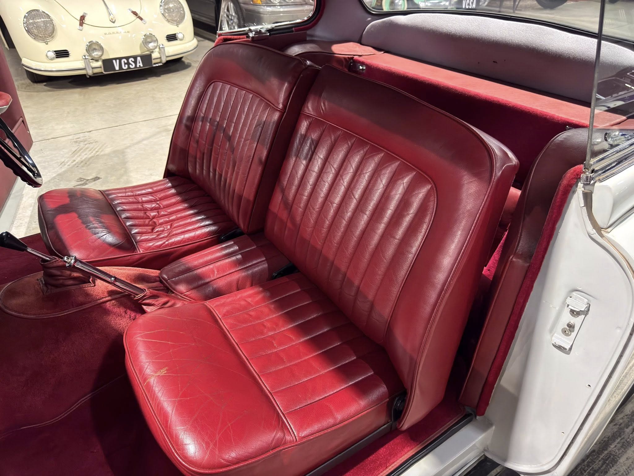 Red leather seats inside a vintage convertible, with burgundy carpeting and chrome trim visible by the doorframe while parked in a showroom.