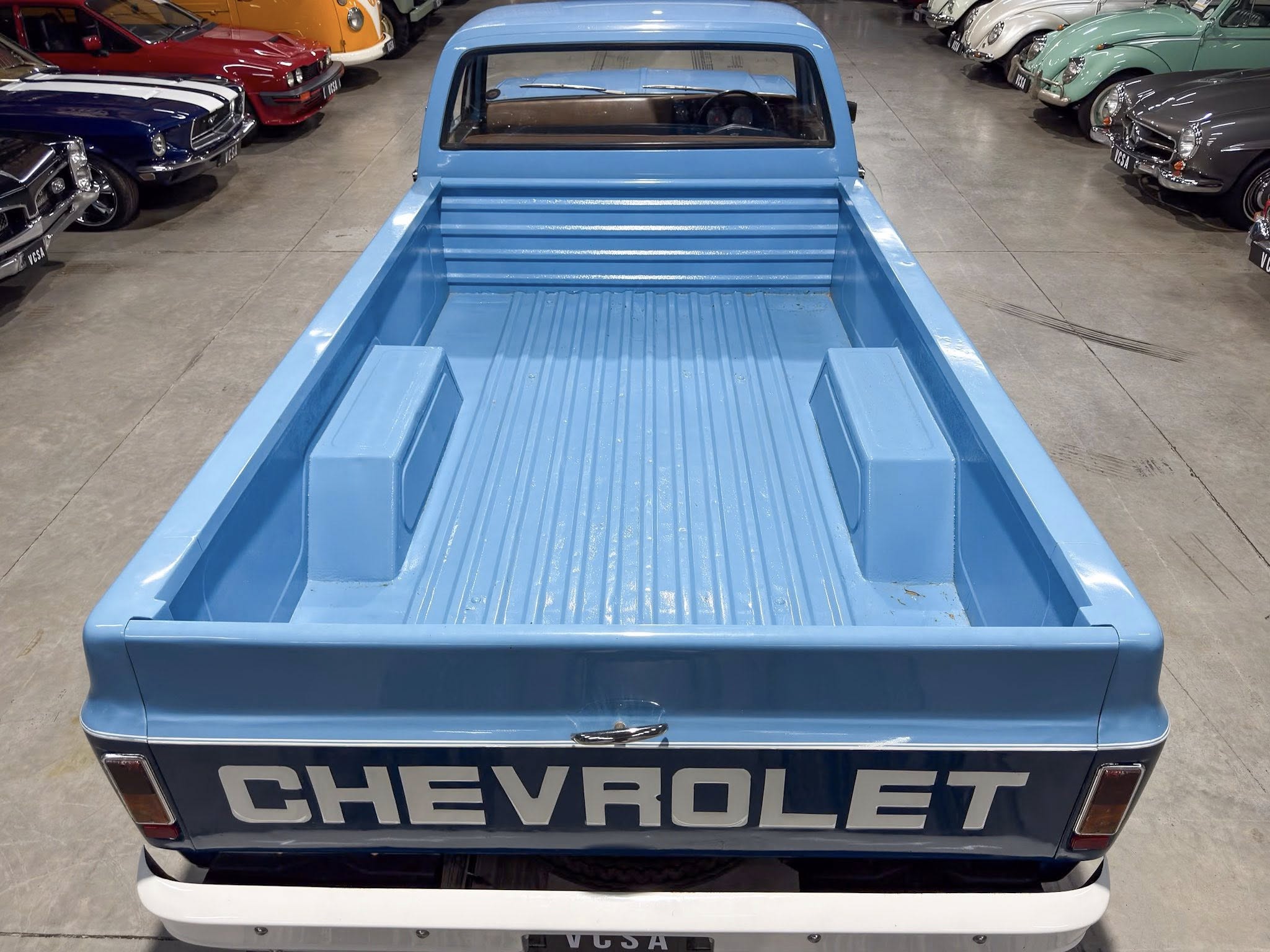 Rear view of a blue vintage Chevrolet pickup in a showroom, bed visible and surrounded by classic cars on both sides.