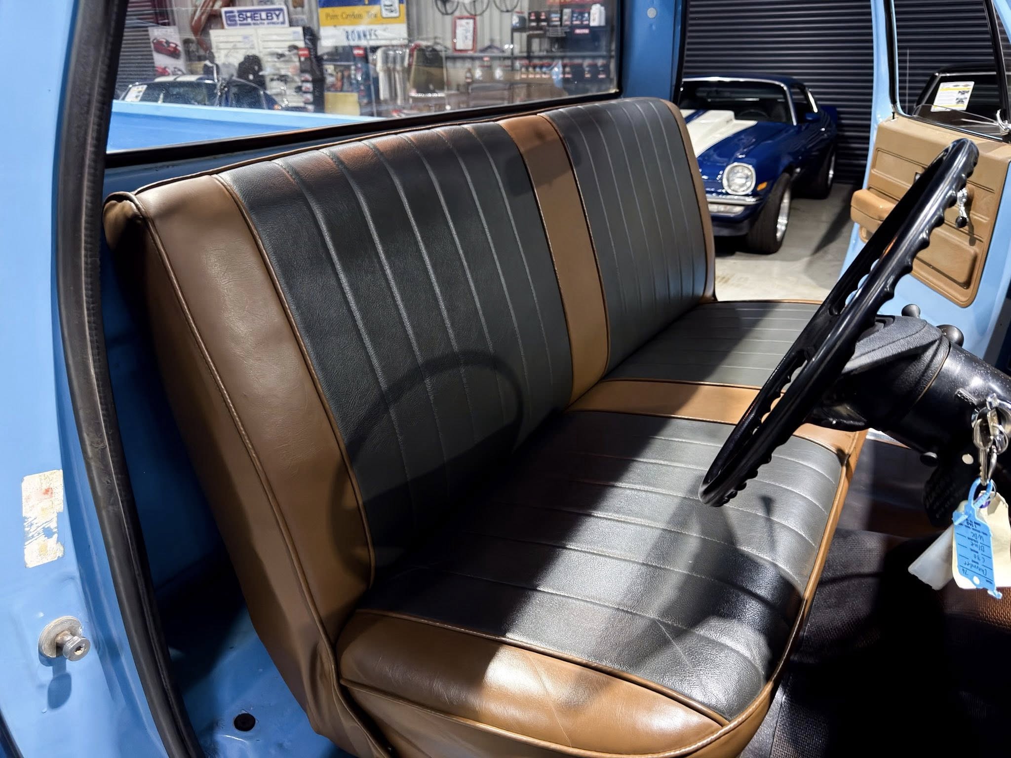 Blue classic car interior with brown and black vinyl bench seat, open door, showroom visible outside the cabin window in the background.