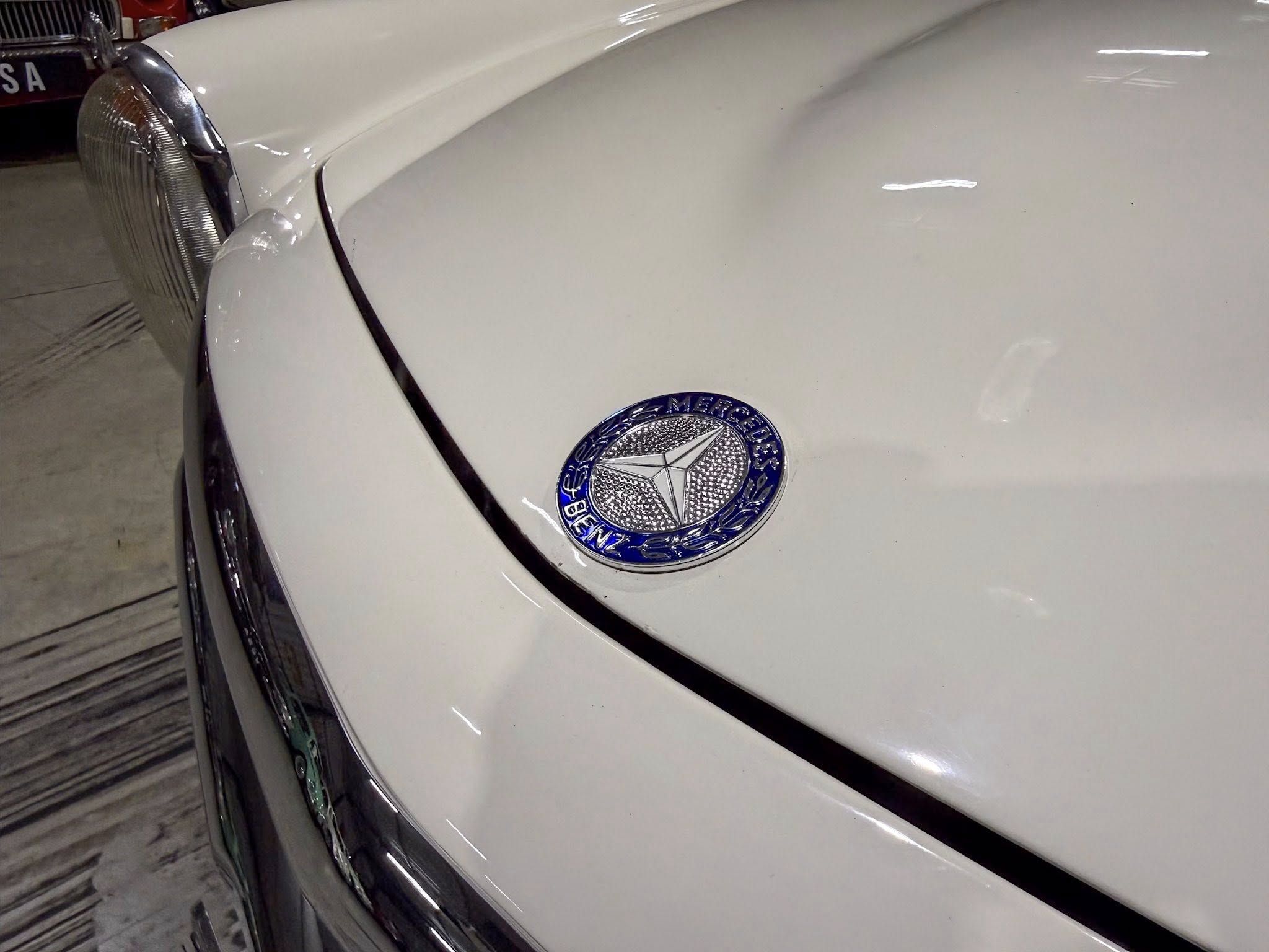 Close-up of a white vintage Mercedes hood with the emblem visible, and the left headlight in a garage setting.