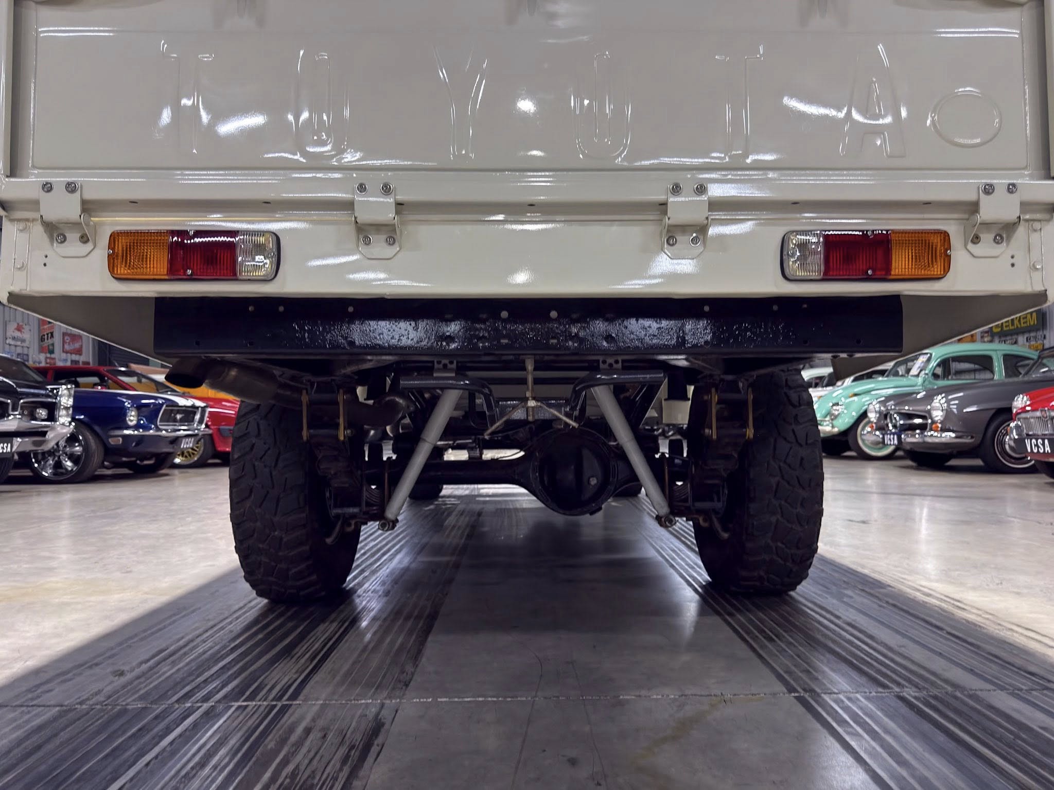 Rear view of a white lifted off-road truck on a showroom lift, displaying large tires and taillights with a row of classic cars in the background.