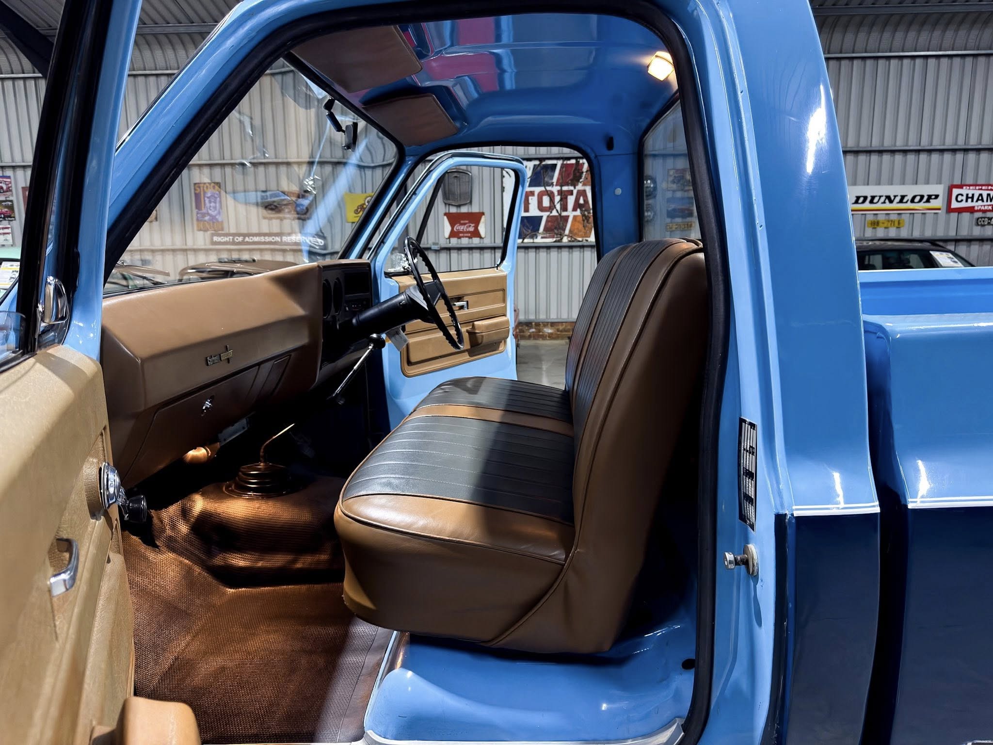 Interior of a blue vintage pickup truck with tan seats, open driver door, and brown dashboard visible in a workshop setting.