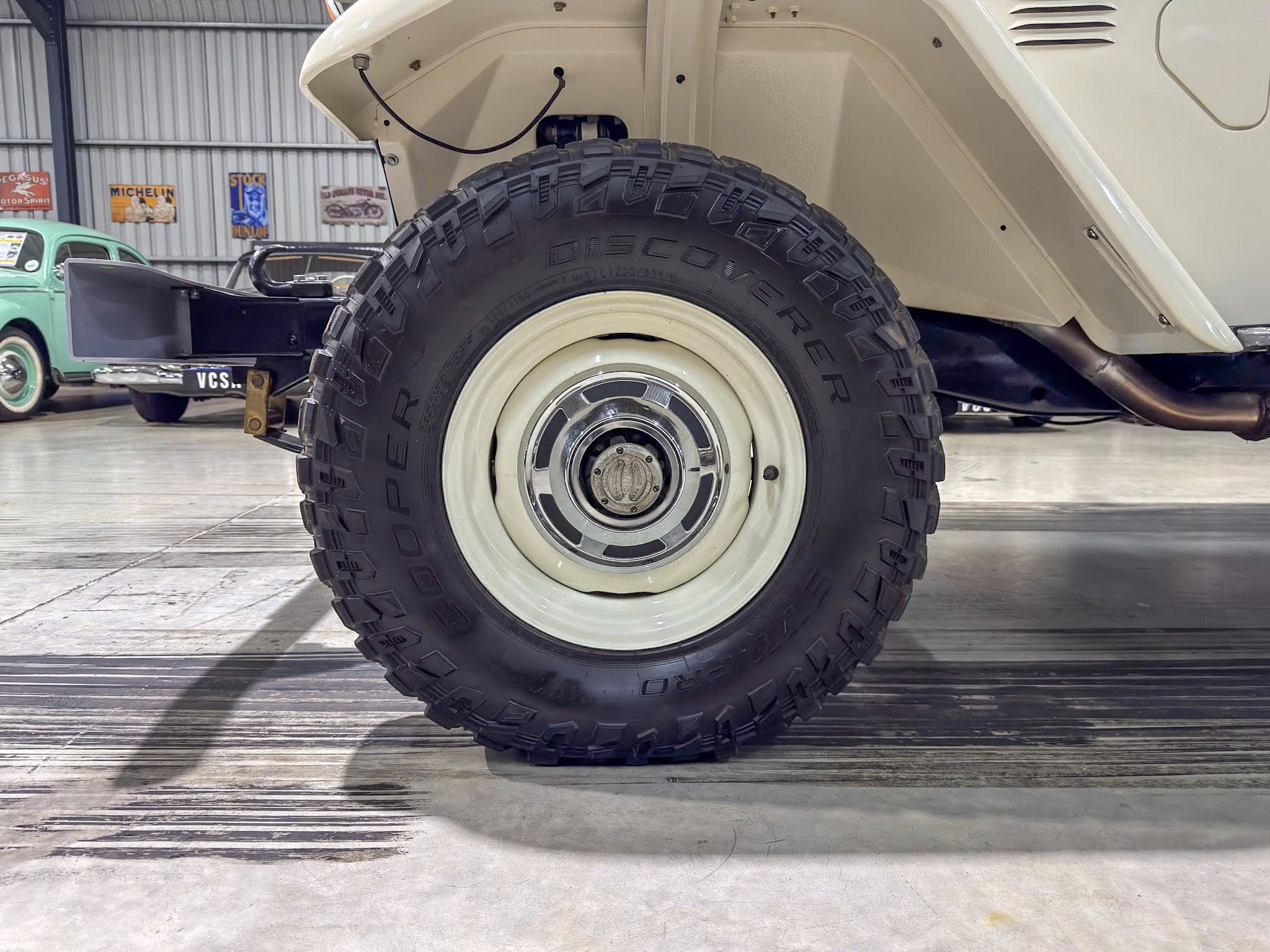 Close-up of a large off-road tire and white wheel under a light-colored vehicle in a garage with vintage posters on the wall behind it.