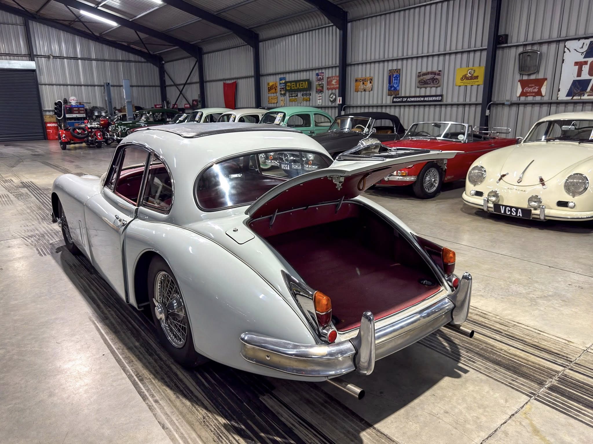 Rear view of a silver vintage car with its trunk open in a showroom filled with more classic cars and retro signage on the walls.