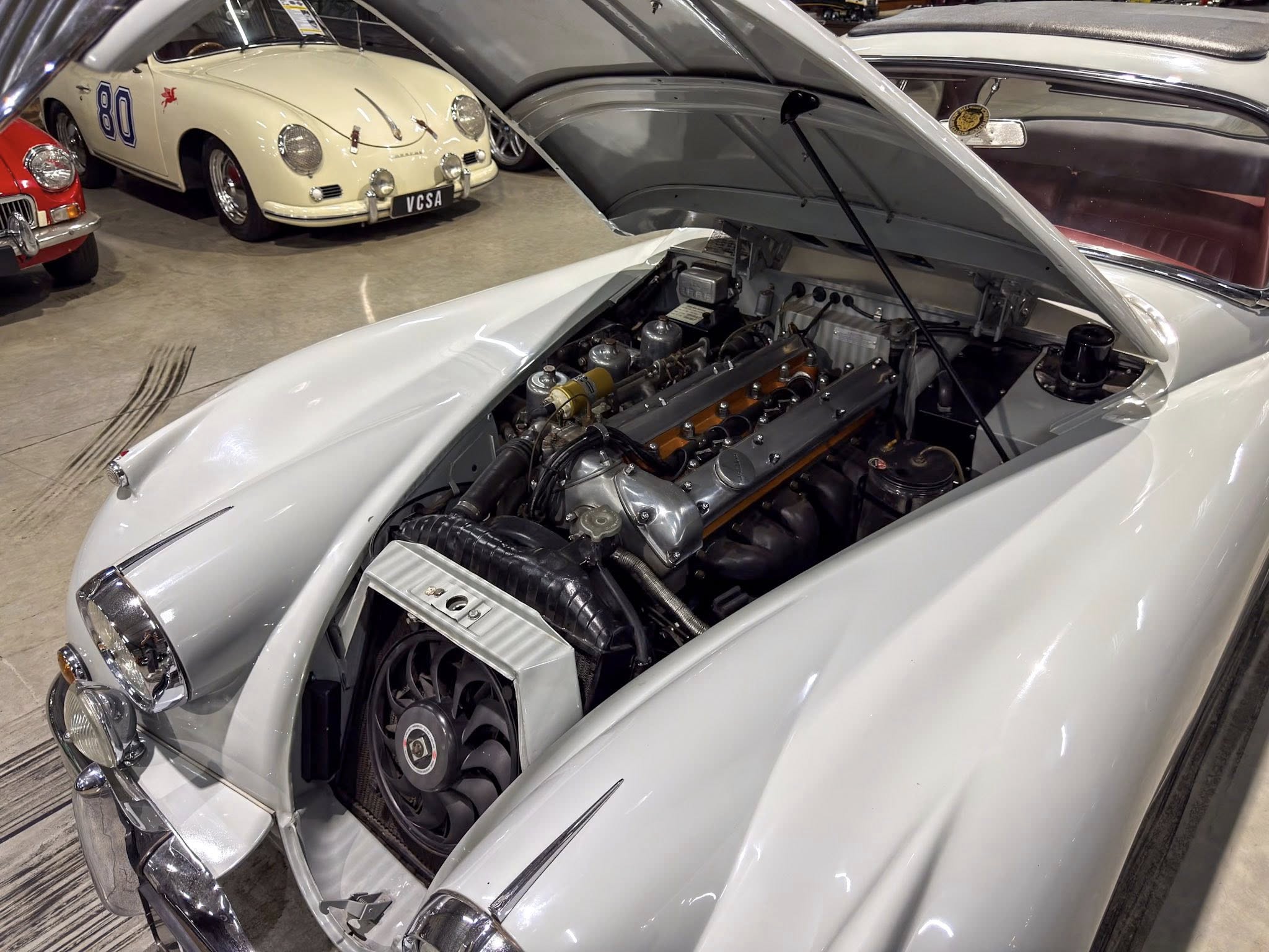 Open car hood exposing classic engine bay of a silver vintage sports car in a showroom with other vintage cars in the background