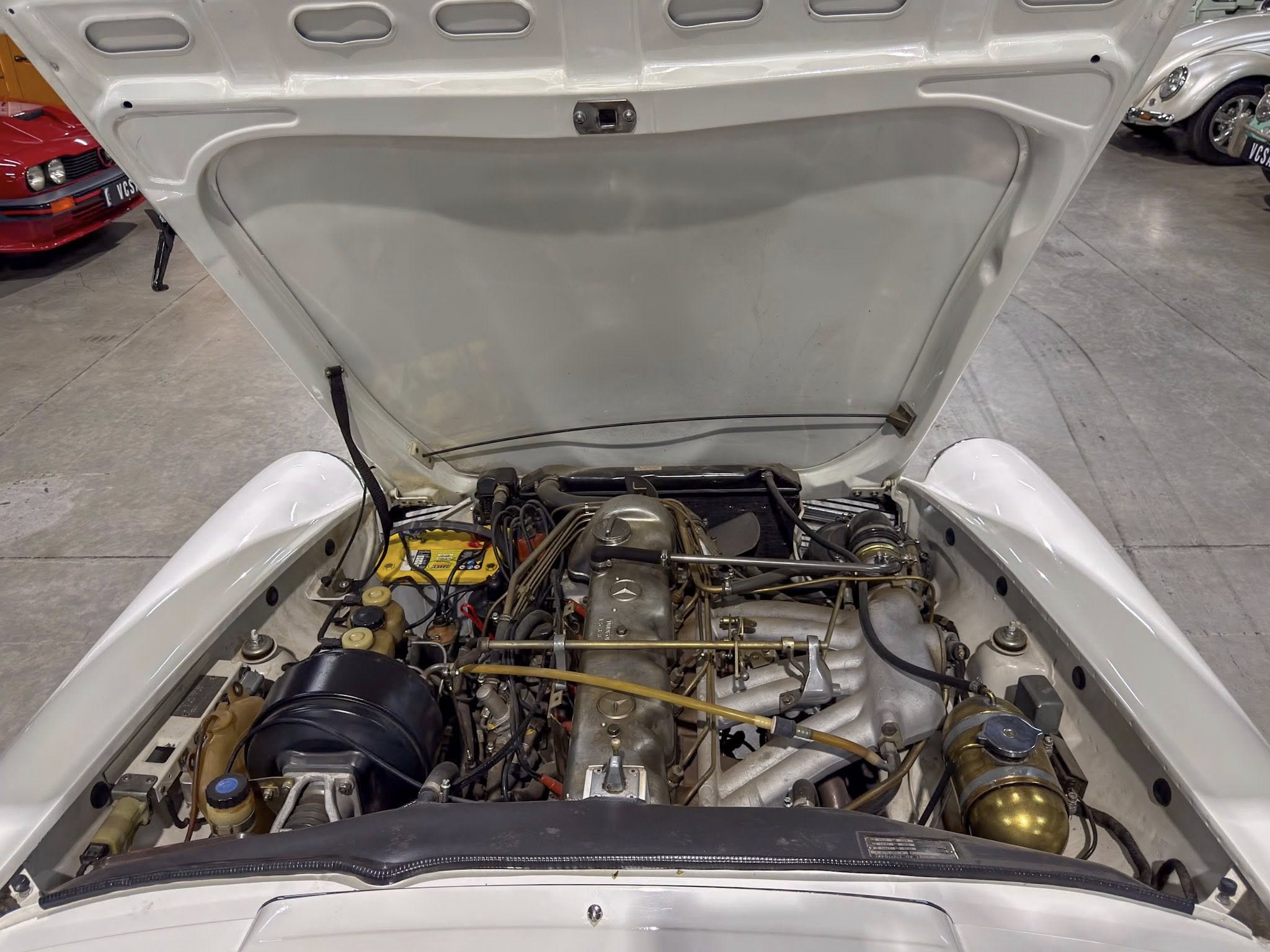 Open hood view of a classic car engine bay with exposed mechanical components and a yellow battery visible on the left side in a showroom setting