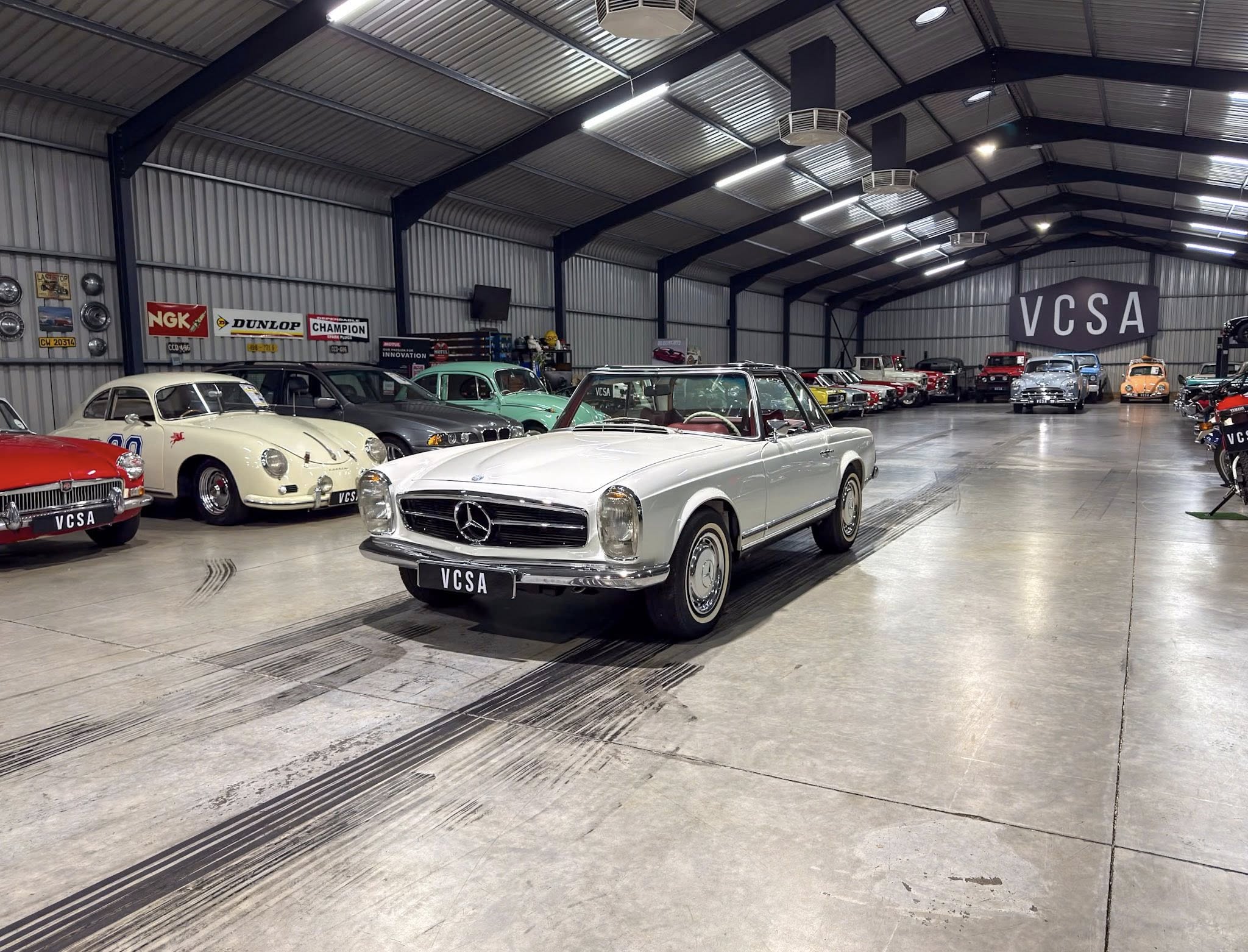 Showroom full of vintage cars on display, with a prominent VCSA banner in the back and a silver Mercedes at the front center.