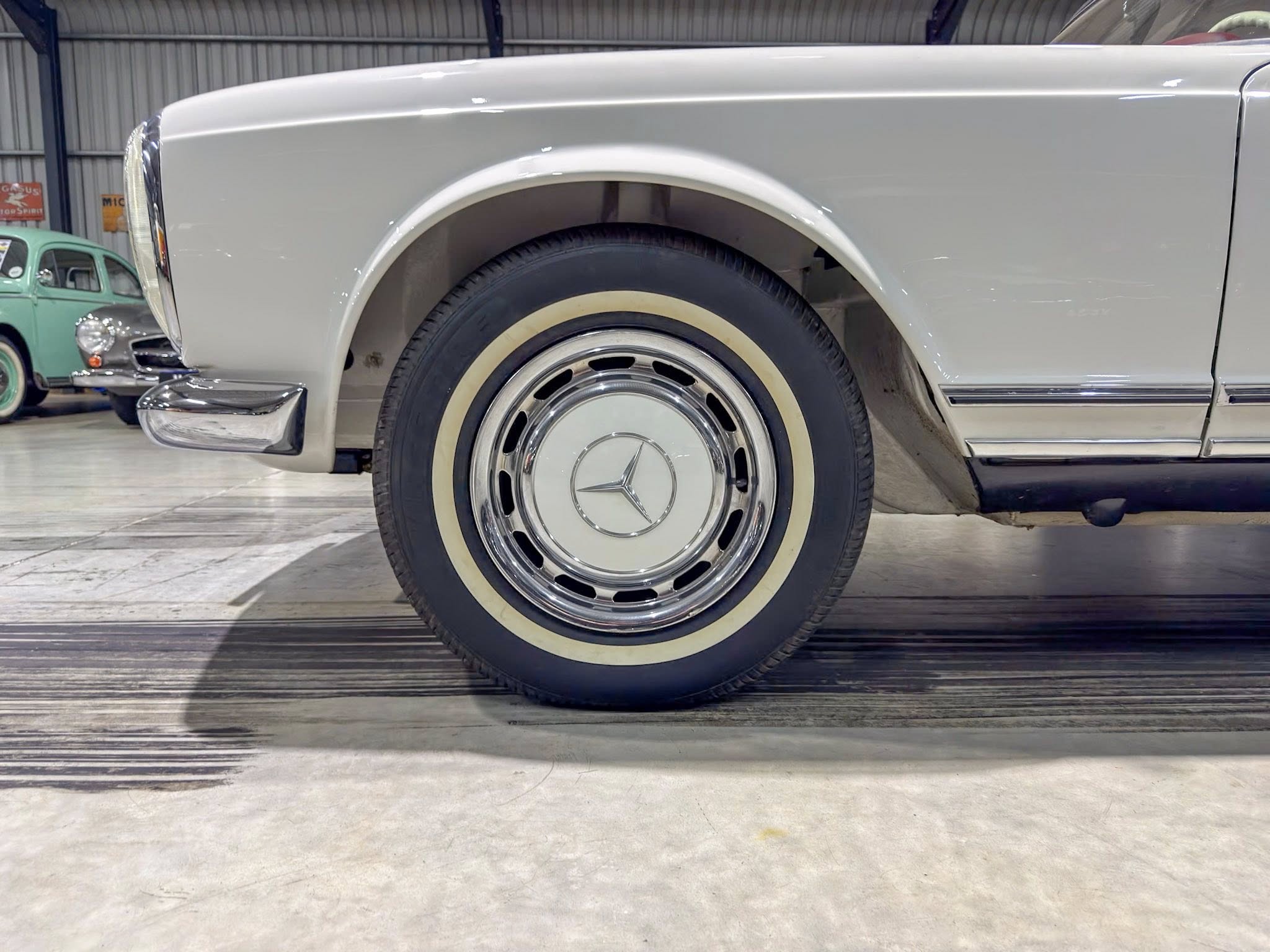 Close-up of a vintage car wheel: cream-whitewall tire with a chrome hubcap and Mercedes-Benz star emblem in a showroom setting.