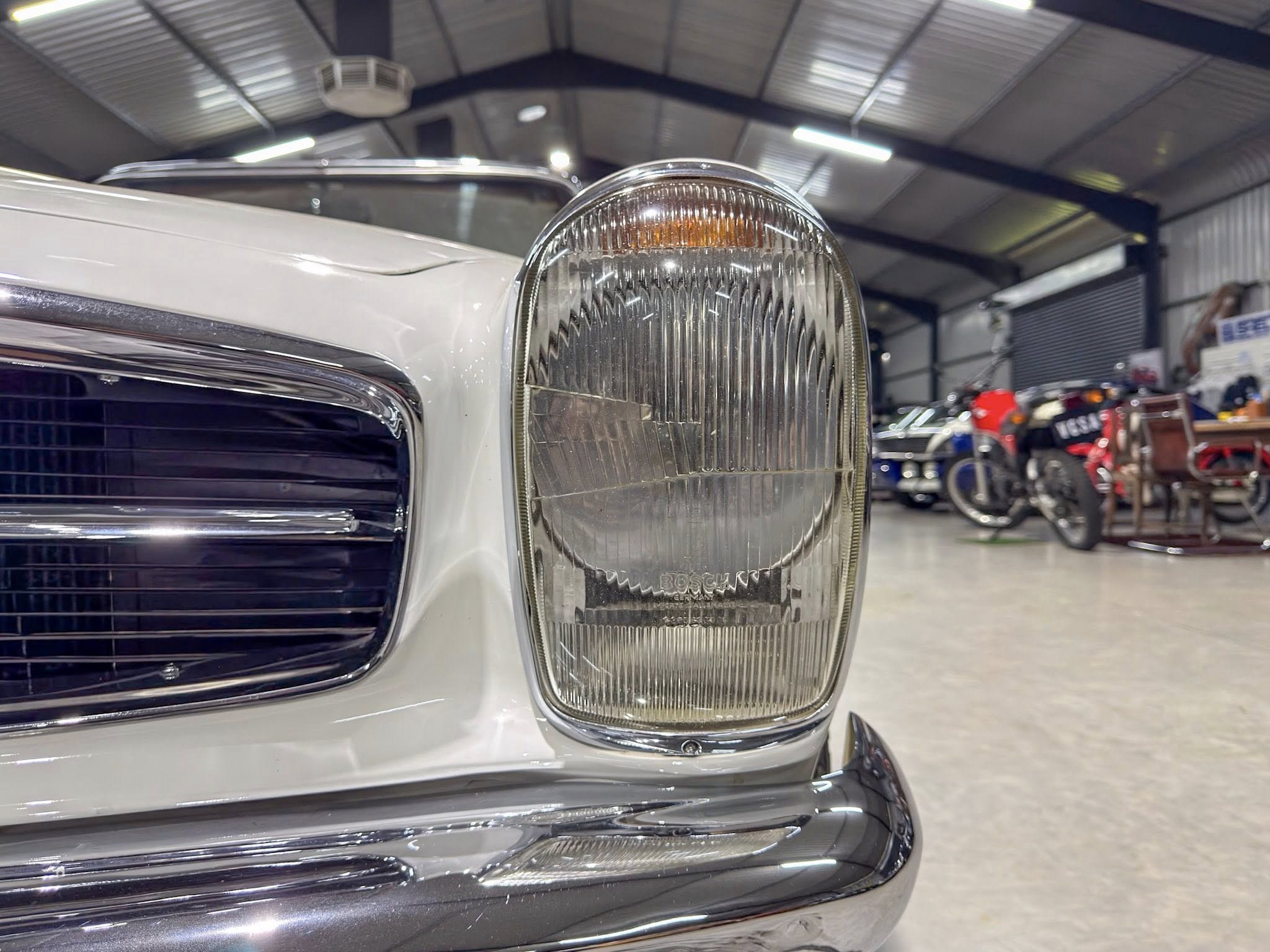 Close-up of a vintage car headlight and chrome grille inside a showroom with other cars in the background
