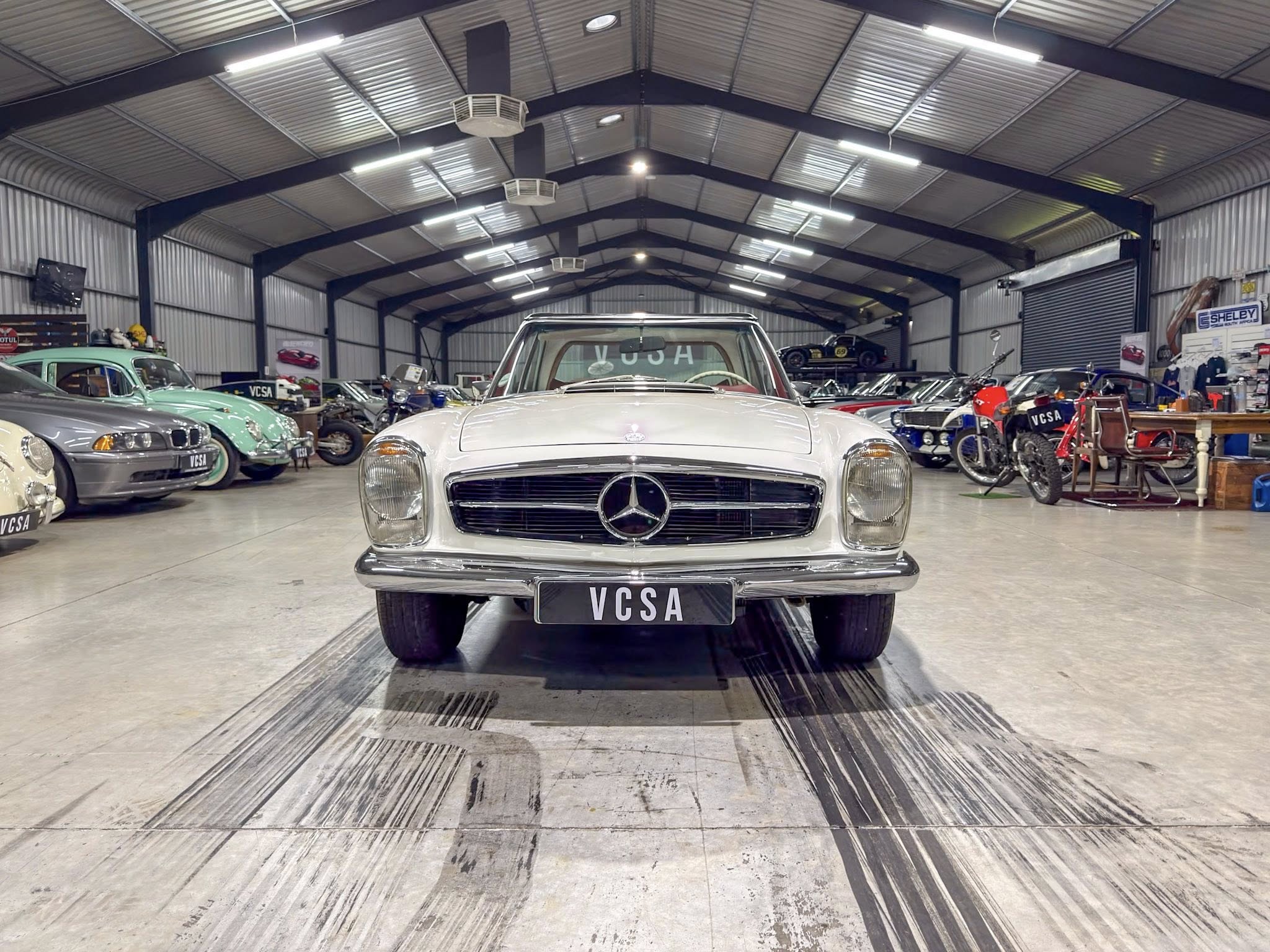 Front view of a silver vintage Mercedes in a busy car showroom with other classic vehicles around it.