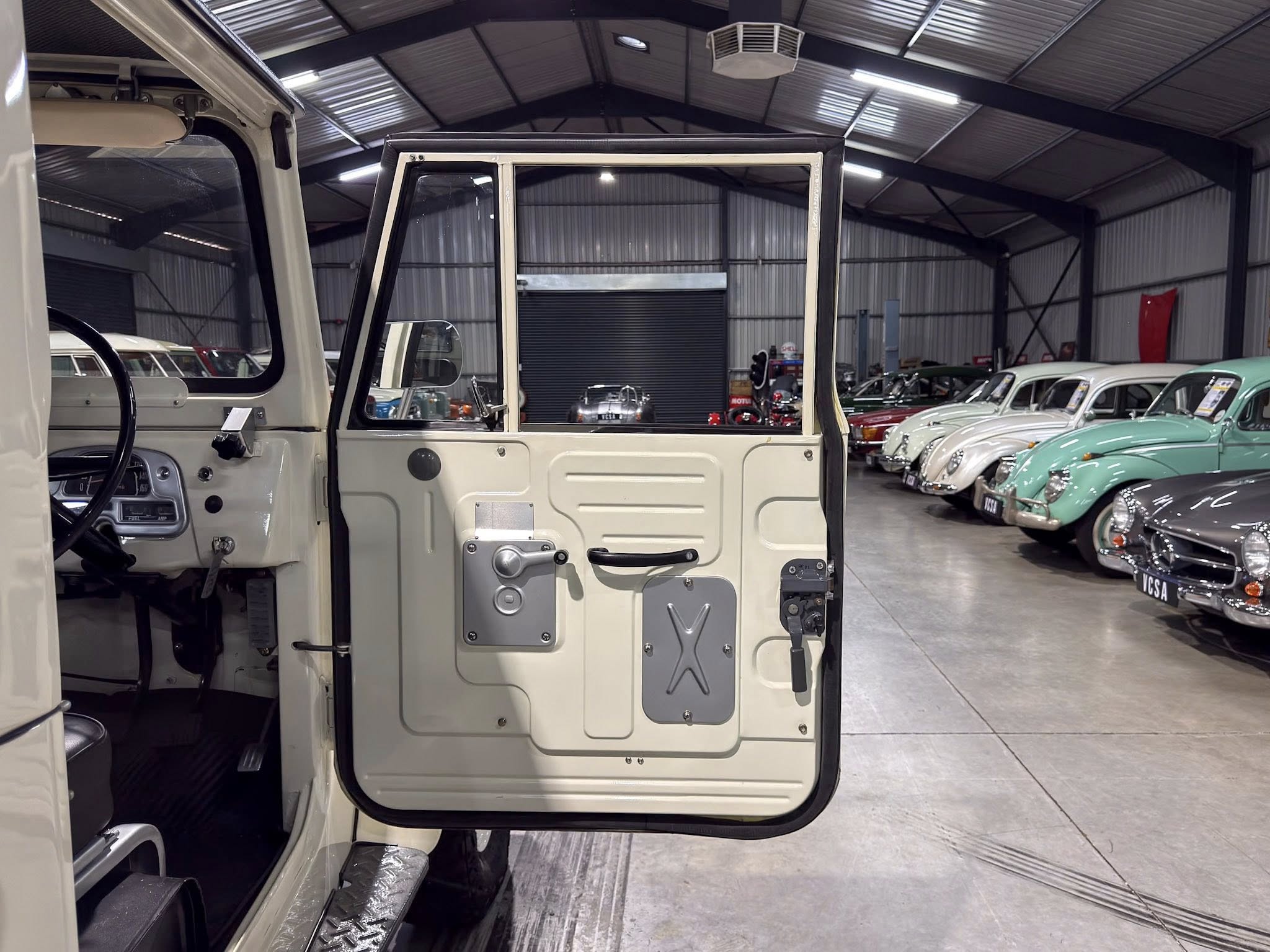 Open beige vintage car door revealing its interior, with a row of classic cars parked inside a showroom.