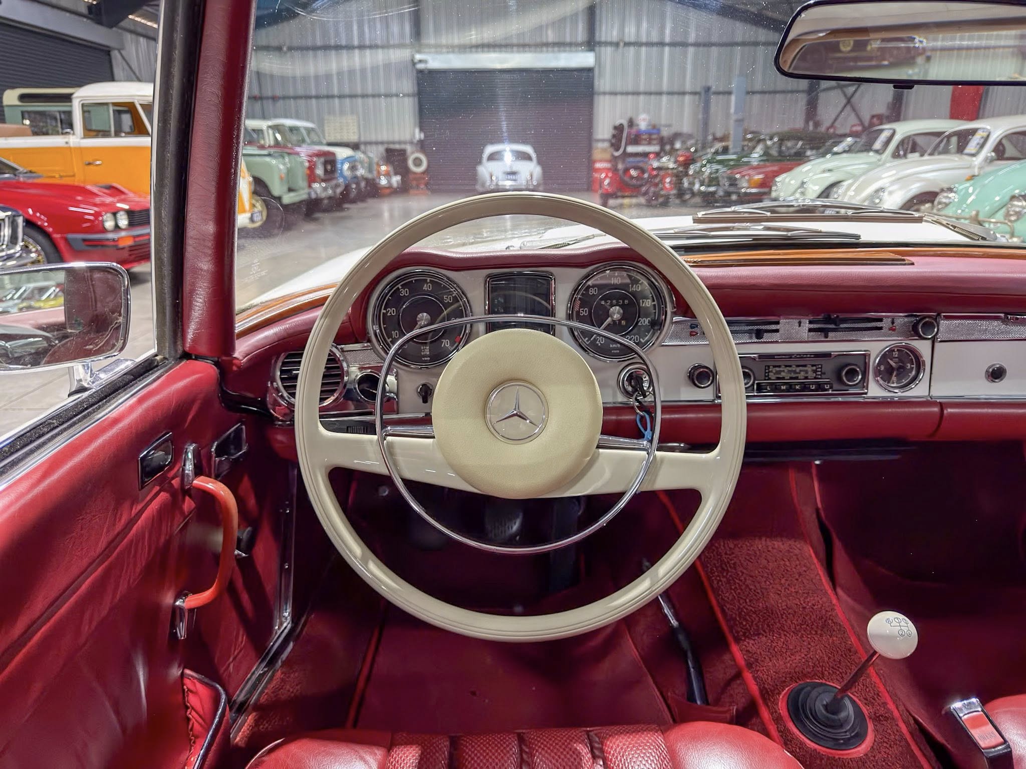 View from the driver's seat of a red vintage car, showing a cream steering wheel and retro dashboard with a showroom of classic cars in the background.