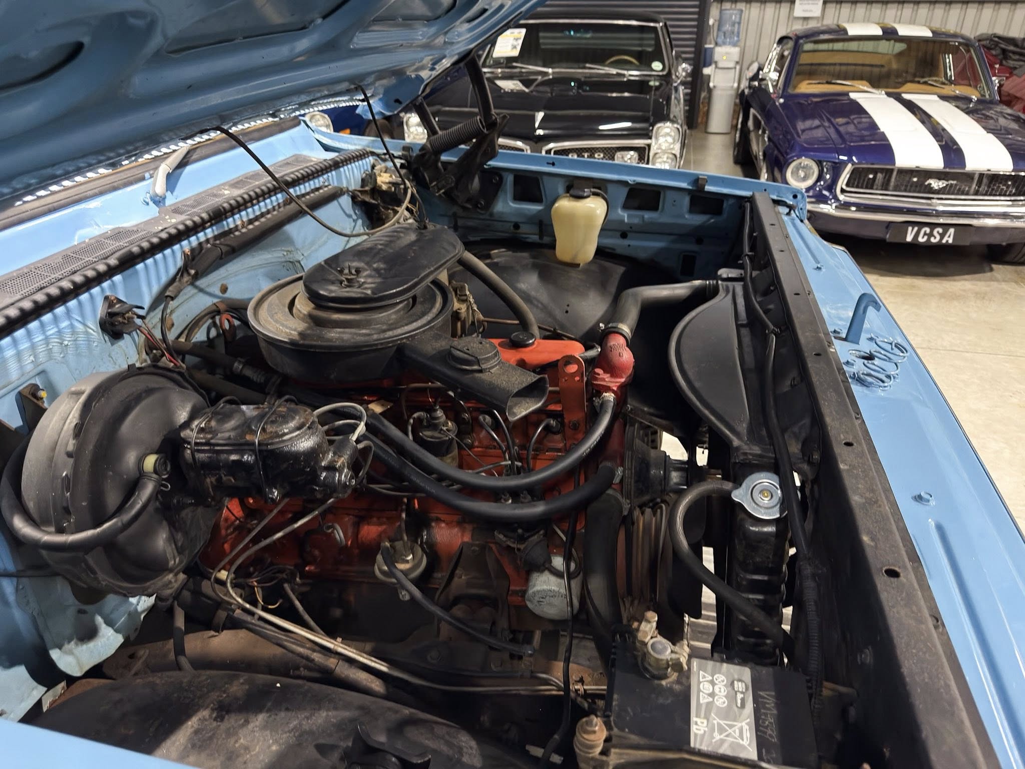 Open hood of a blue car showing its engine bay with hoses and components in a workshop environment