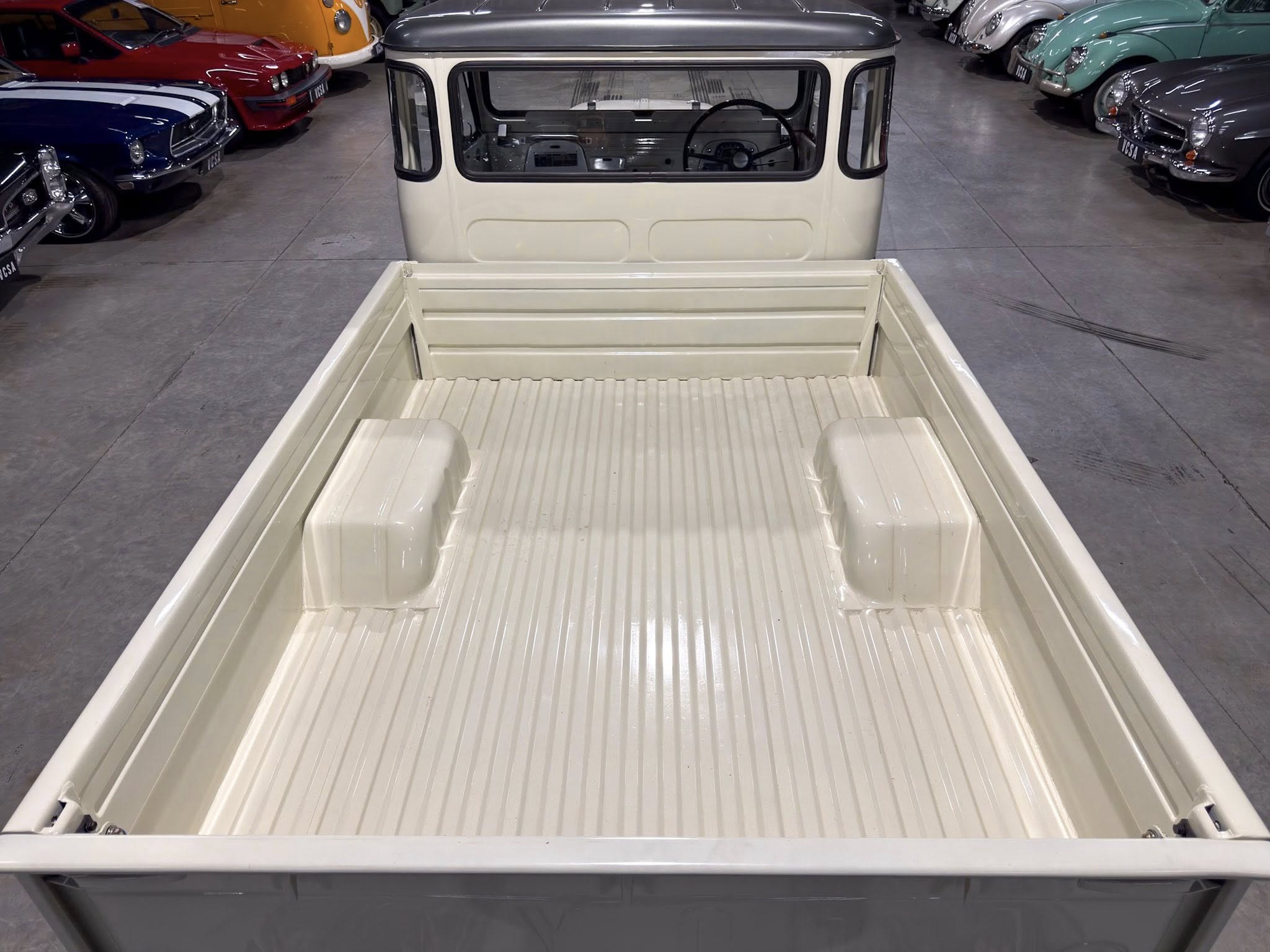 Rear of a cream pickup truck with a ribbed bed in a classic-car showroom, viewed from the back with vintage cars on both sides.