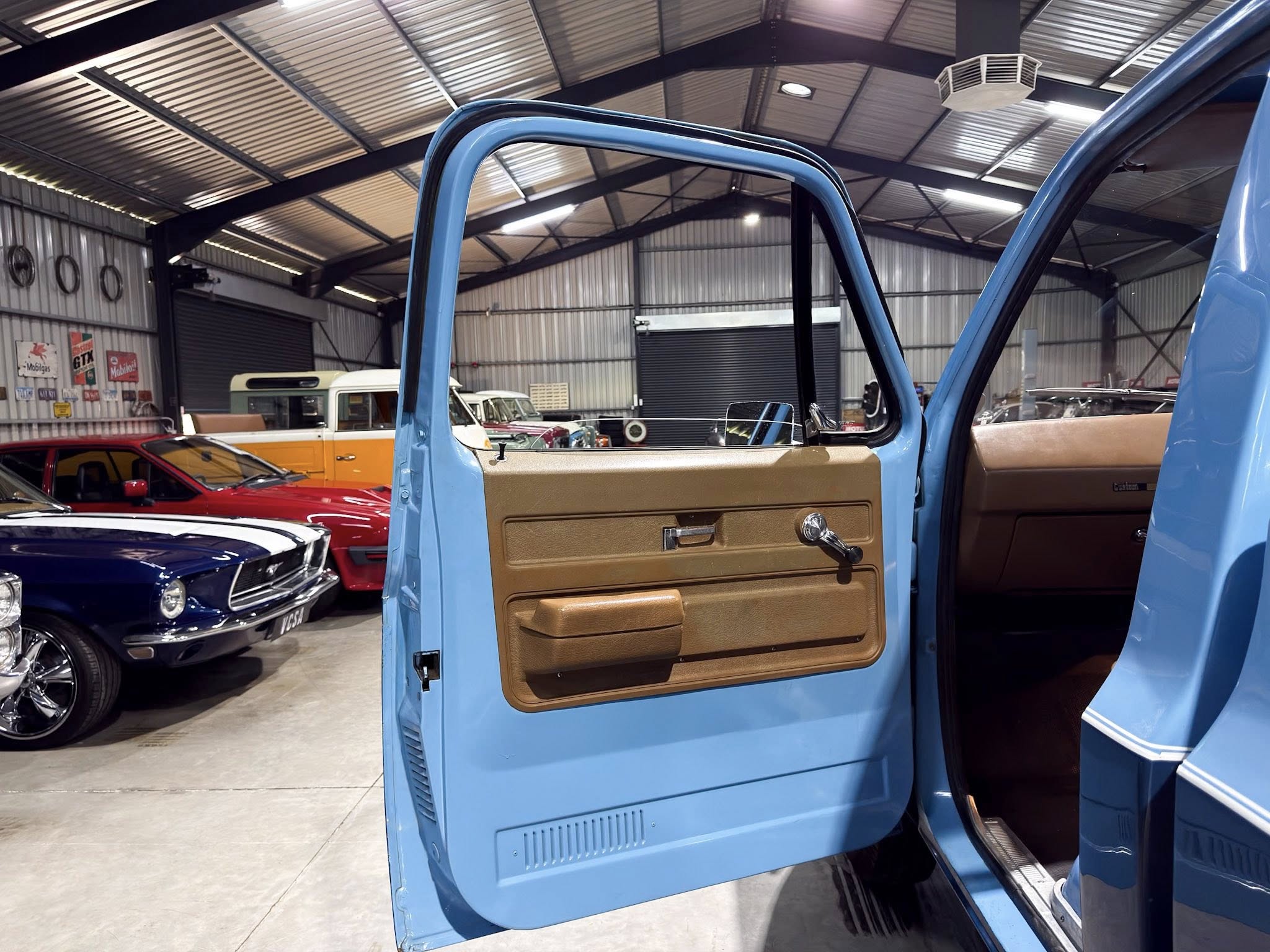 Open blue vintage vehicle door with tan interior inside a classic car showroom or garage, with other vintage cars visible in the background.