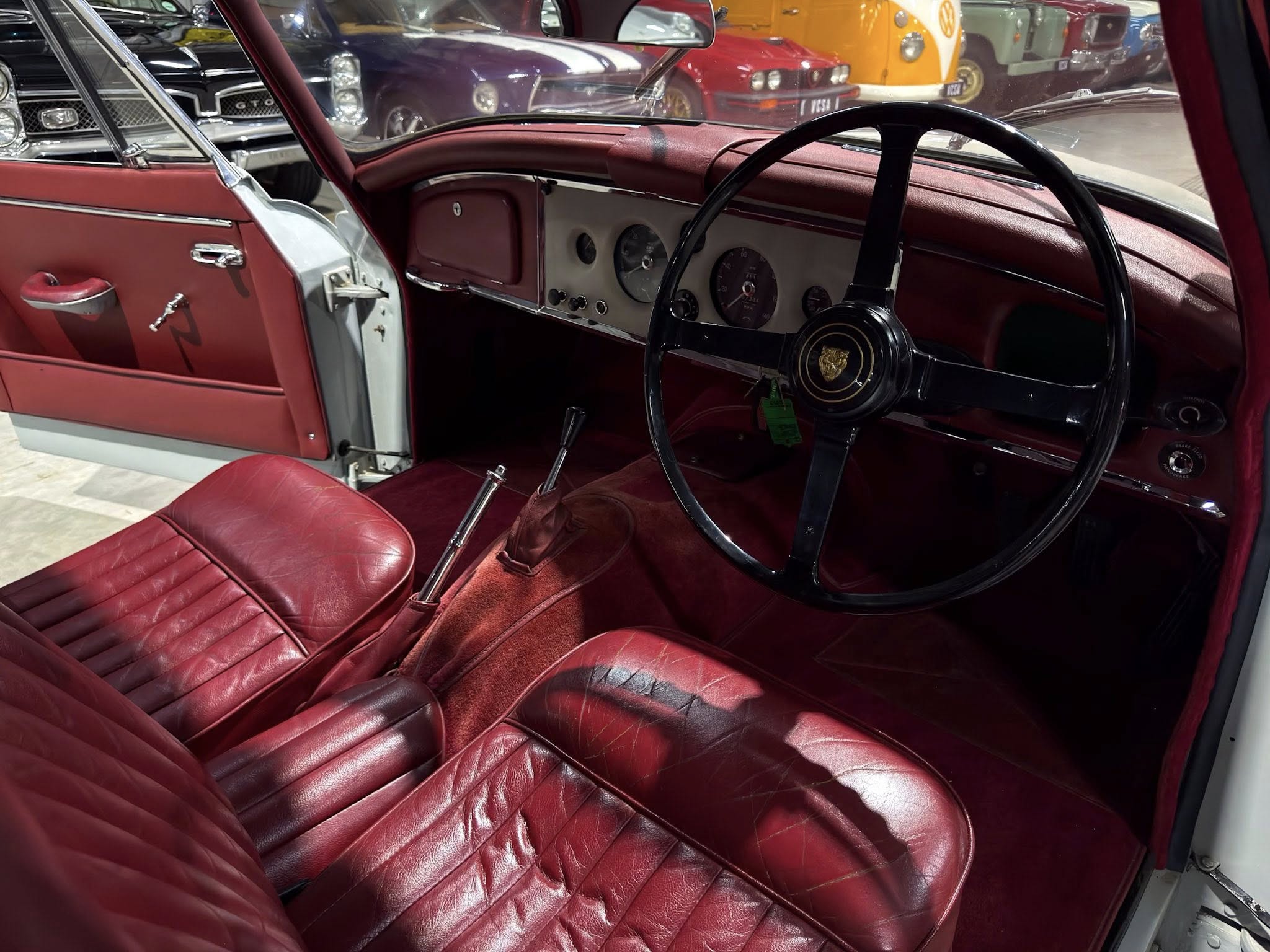 Red vintage car interior with red leather seats, chrome accents, and a black steering wheel; other classic cars visible through the window.