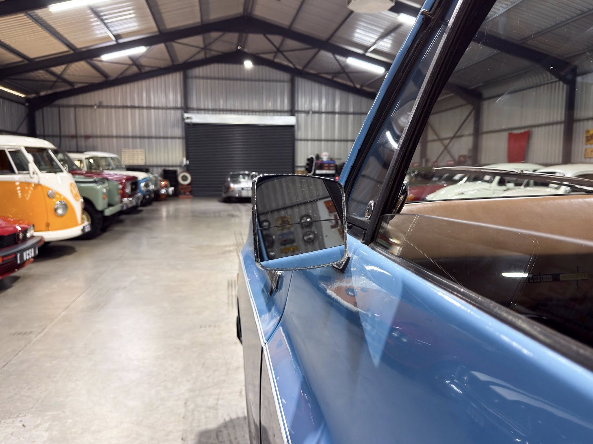 Blue vintage car close-up with side mirror in a large indoor showroom filled with colorful classic cars along the left, metal beam ceiling overhead.