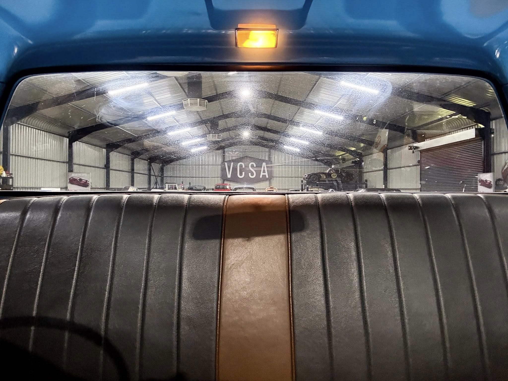 View from the back seat of a car, looking through the rear window into a large garage with parked cars and a banner reading 'VCSA' in the distance; blue exterior paint above the window and black leather upholstery in the foreground.