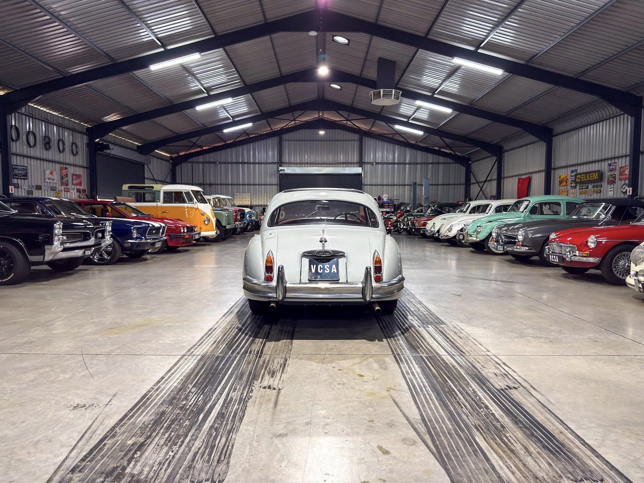 Interior of a vintage car showroom in a large metal warehouse, rows of classic cars on both sides with a white car centered at the back