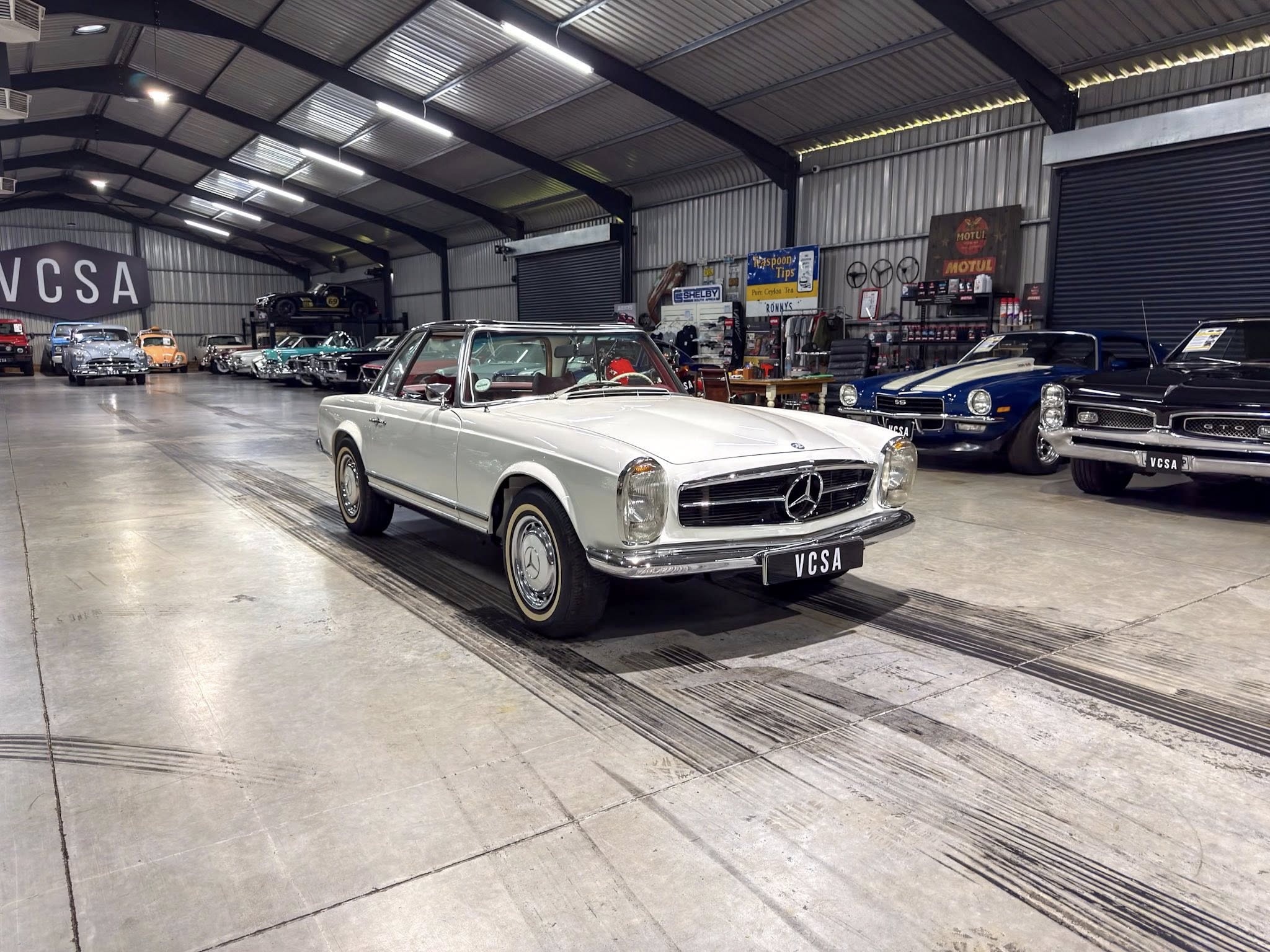 Vintage car showroom inside a large warehouse, with classic convertibles and other retro cars on display; white Mercedes in the foreground.