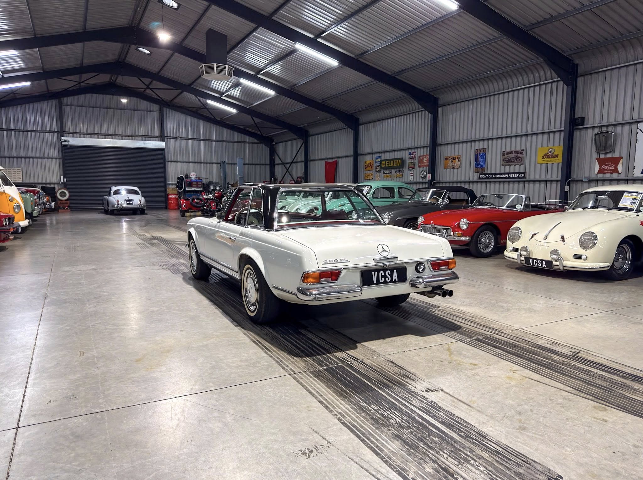Indoor showroom of a vintage car collection with a white convertible in the foreground and classic cars parked along the right wall inside a metal warehouse.