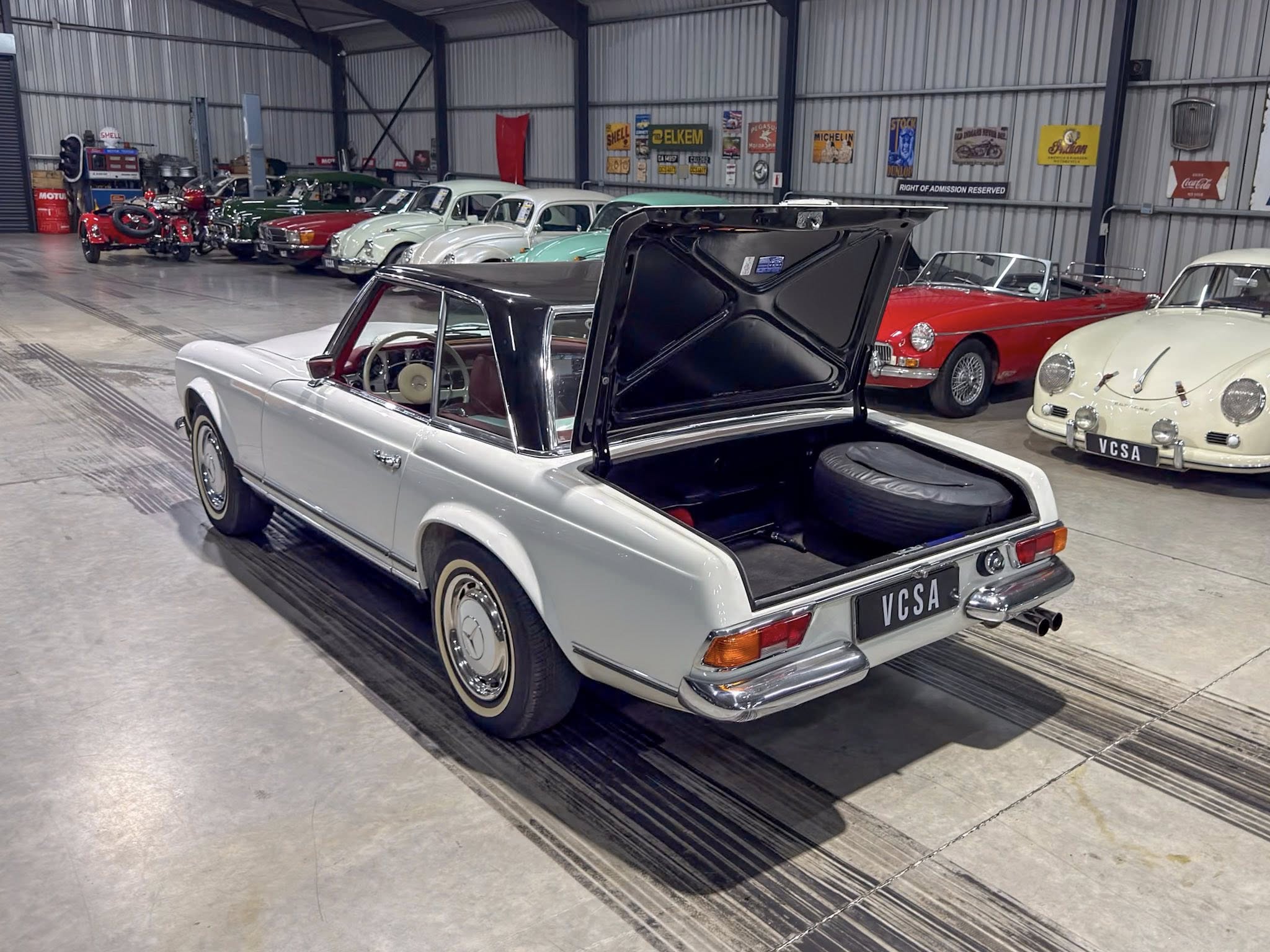 Line of vintage cars inside a spacious garage or showroom, with a silver convertible in the foreground and other classic sedans and roadsters parked along the wall behind it, some with pastel colors; one car's trunk is open.